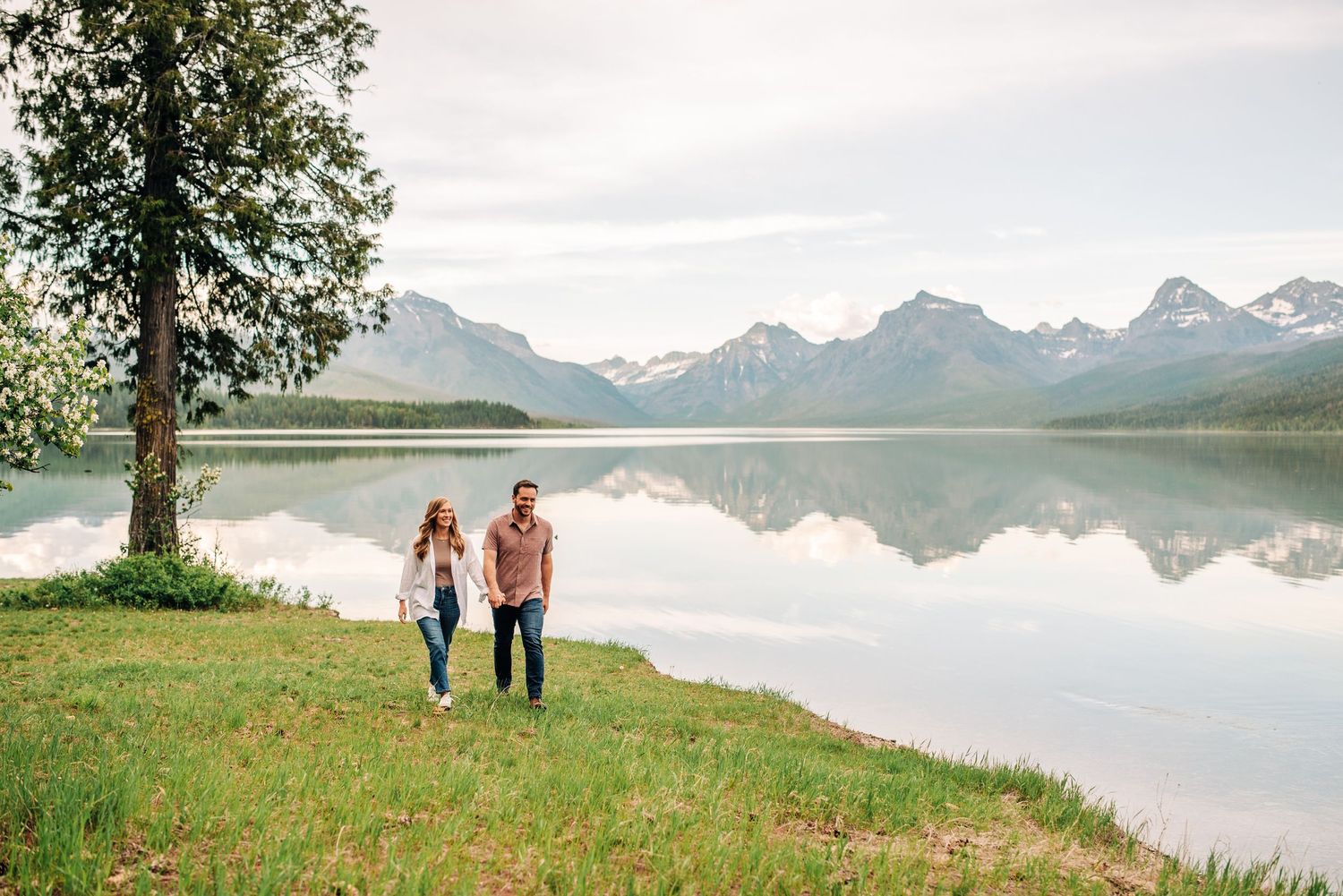 Two people walk hand in hand along a serene mountain lake shore with snow-capped peaks reflected in the calm water.