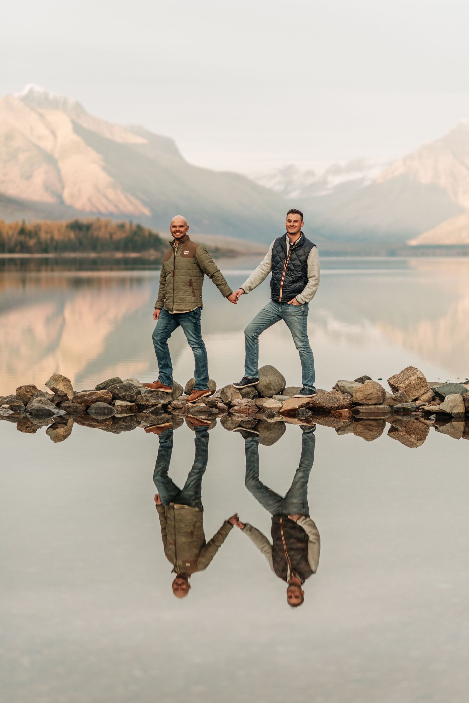 Two people stand on rocks at the edge of a calm mountain lake with perfect reflections at sunset.