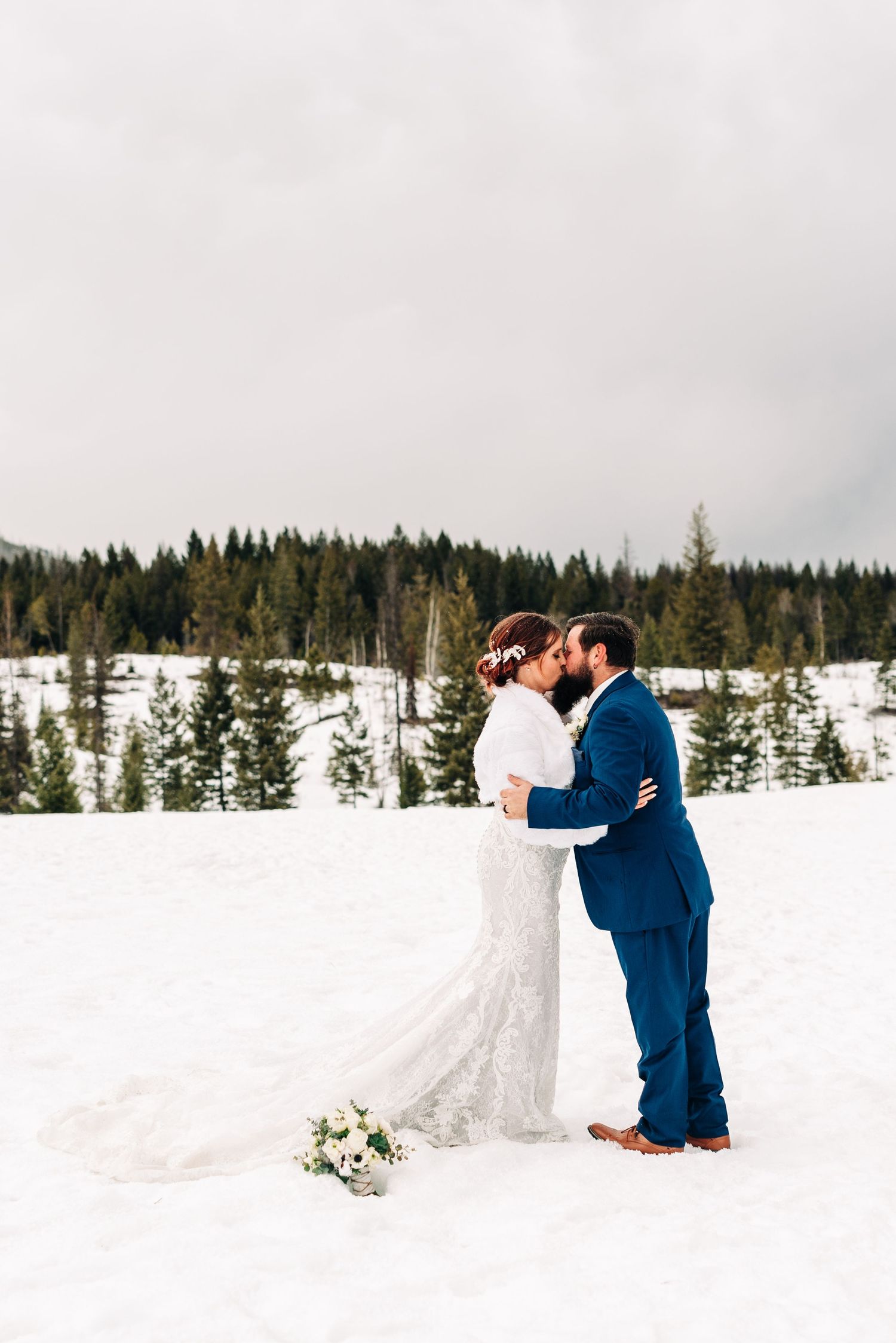 A winter wedding couple embraces in a snowy landscape with evergreen trees in the background.