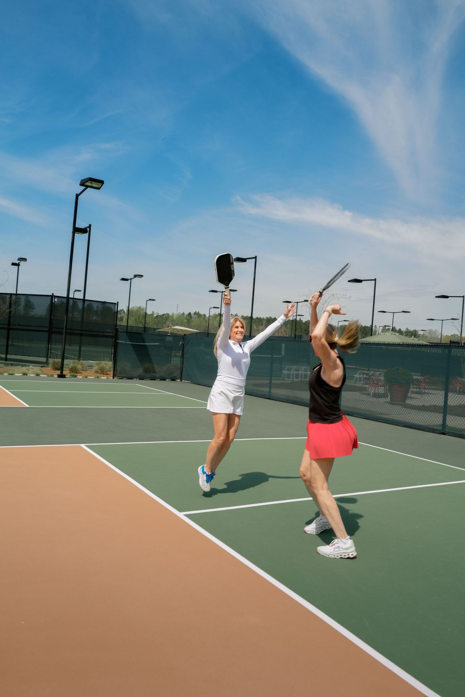 Two tennis players practicing serves on an outdoor court with blue sky and light poles overhead.