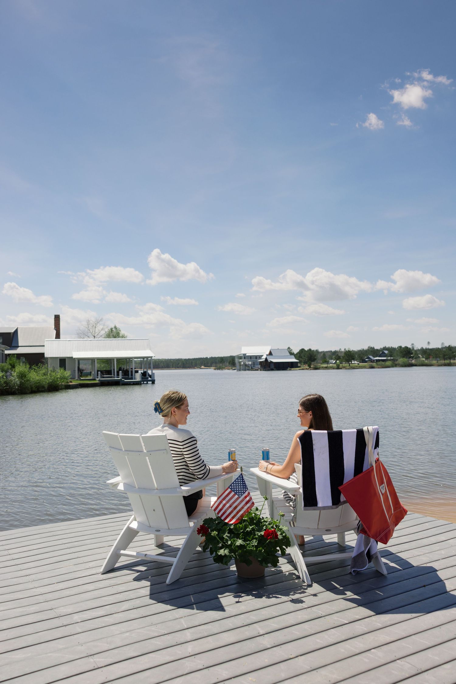 Two people relax in Adirondack chairs on a lakeside dock under a sunny blue sky.