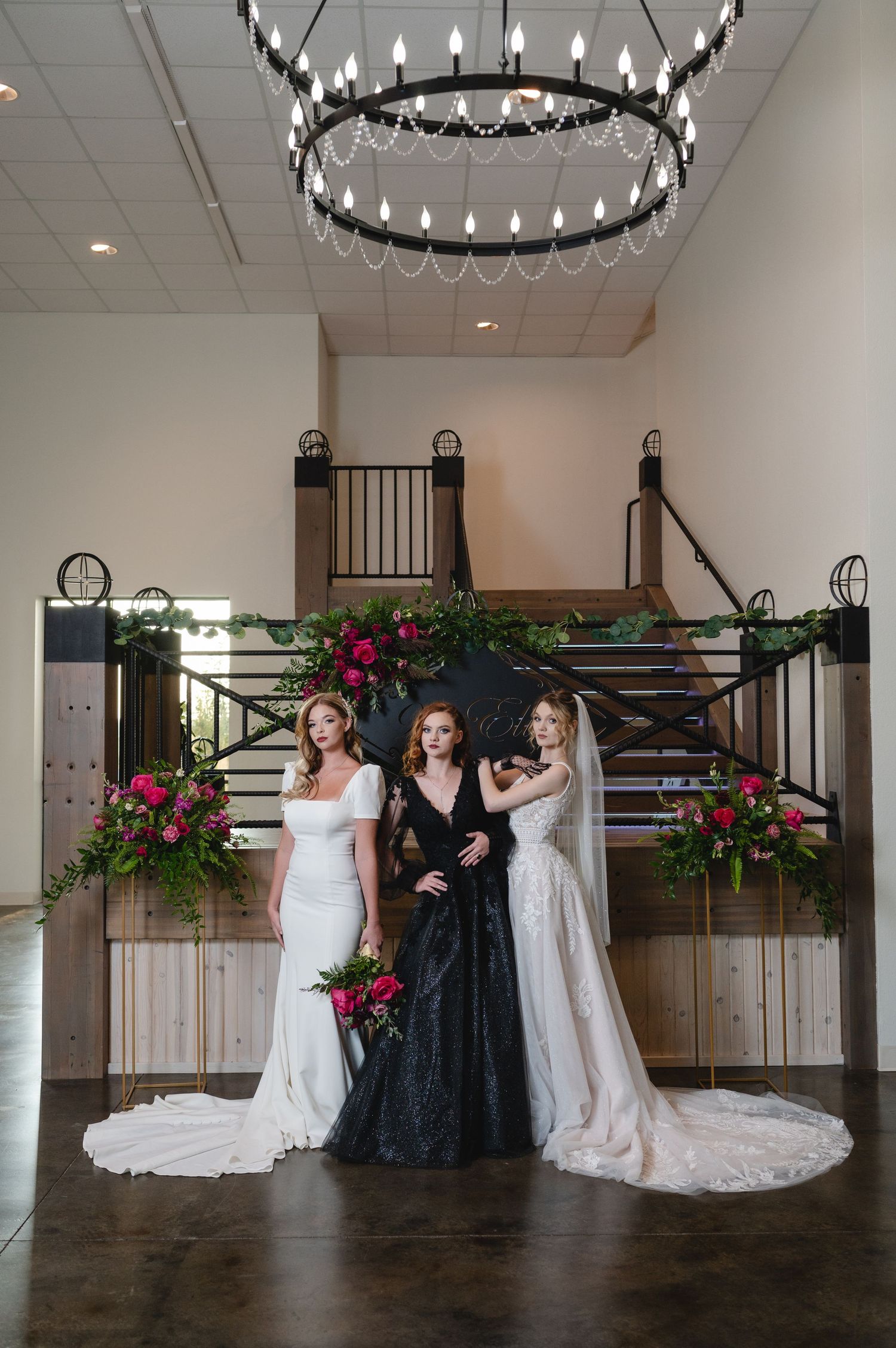 Three people in wedding dresses posing under circular chandelier with floral arrangements.
