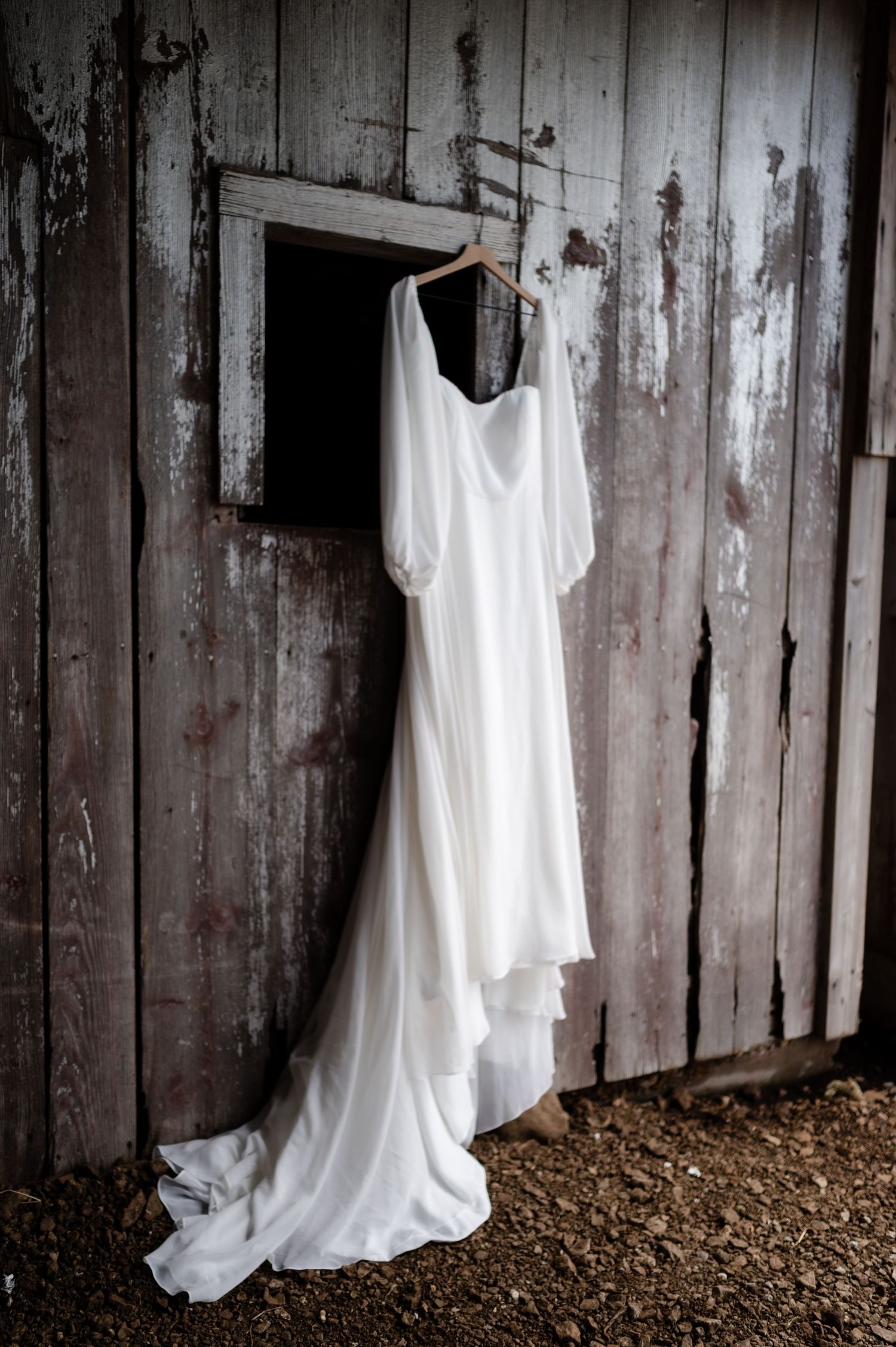 White wedding dress hanging on rustic wooden barn wall in dramatic lighting.