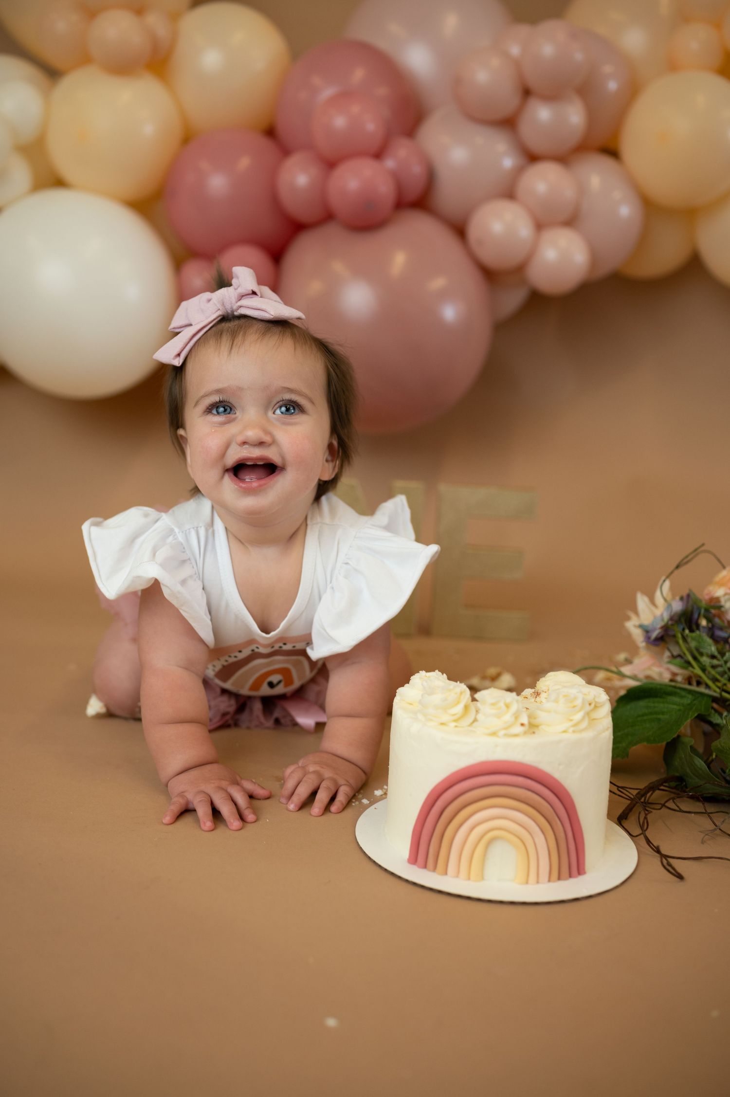 A festive first birthday setup with a rainbow cake and pastel balloon arch against a brown backdrop.