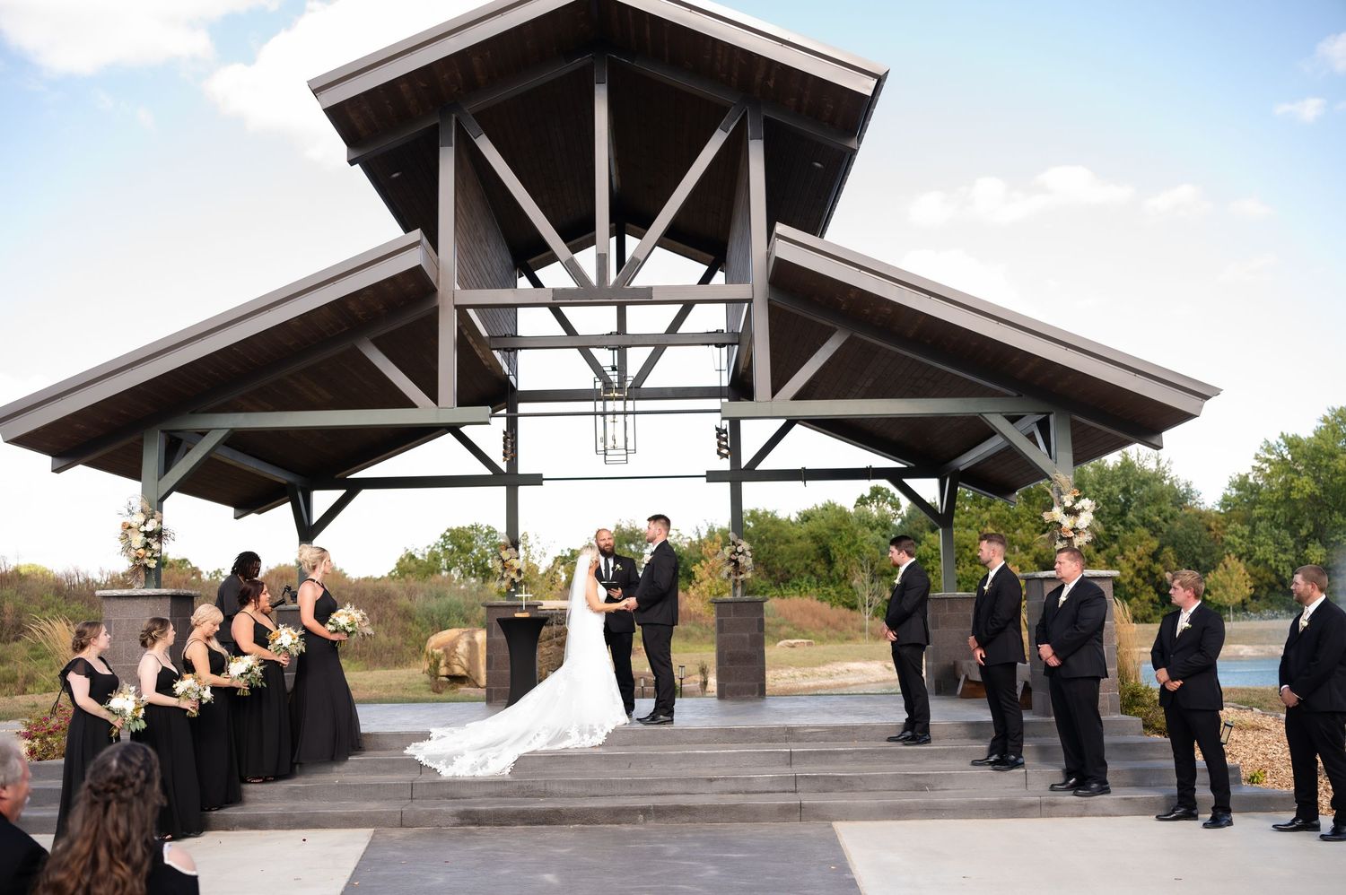 Wedding party poses in front of rustic wooden pavilion with exposed beams during outdoor ceremony.