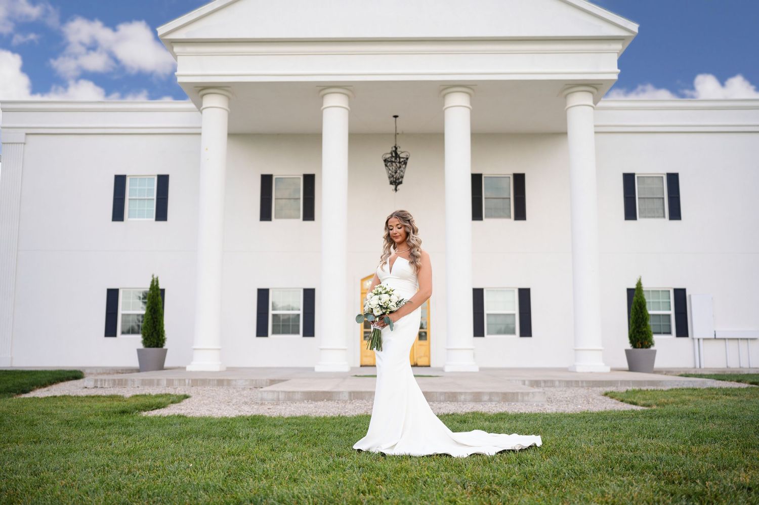 A person in a white wedding dress poses elegantly against a classic white columned building on a sunny day.