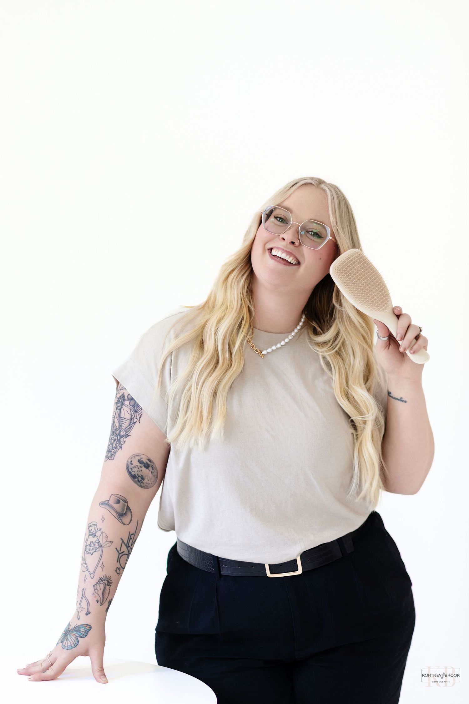 A casual portrait showing tattoos and glasses with a beige top and black pants against white background.