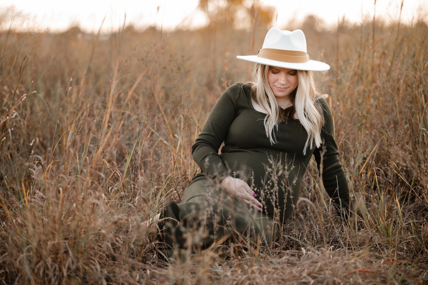 Ethereal maternity portrait in tall grass field during golden hour with woman wearing white hat.