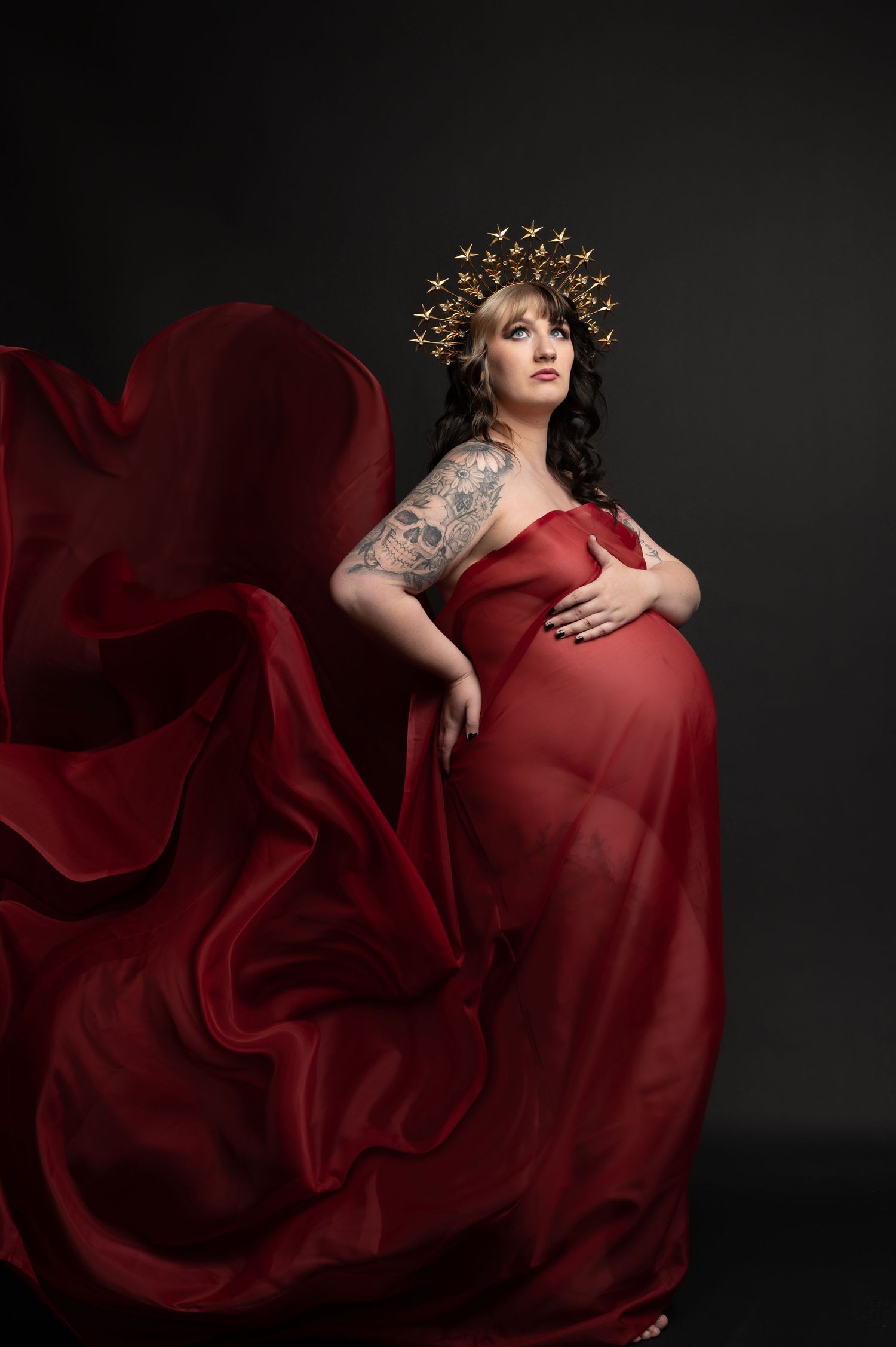 Artistic maternity photo featuring billowing red gown and ornate gold crown against black backdrop.