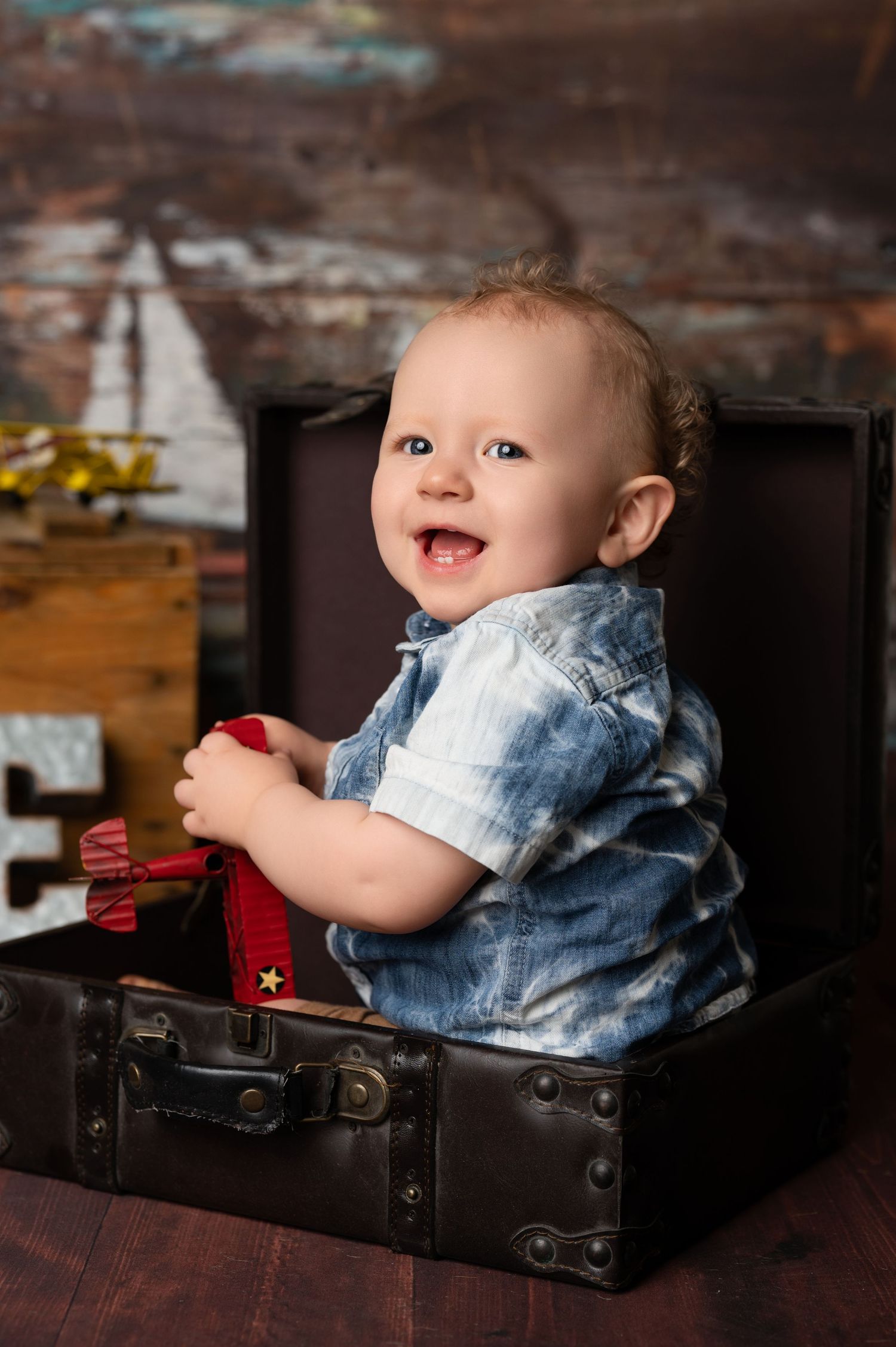A baby in denim clothing smiles while lying in a vintage leather suitcase during a photo session.