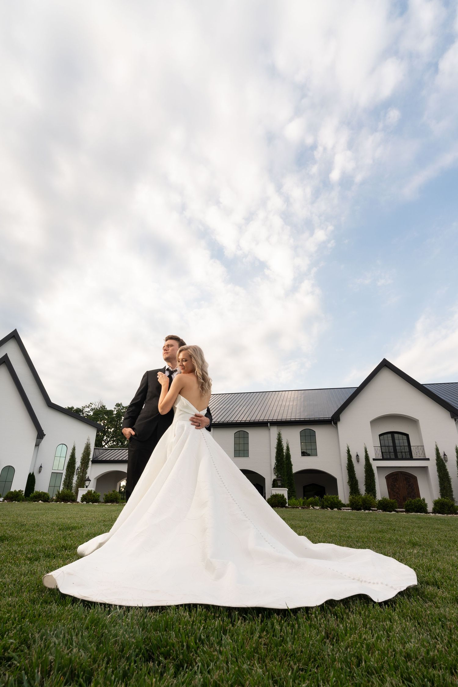 A romantic wedding couple poses together on a lush green lawn with white church buildings in the background.