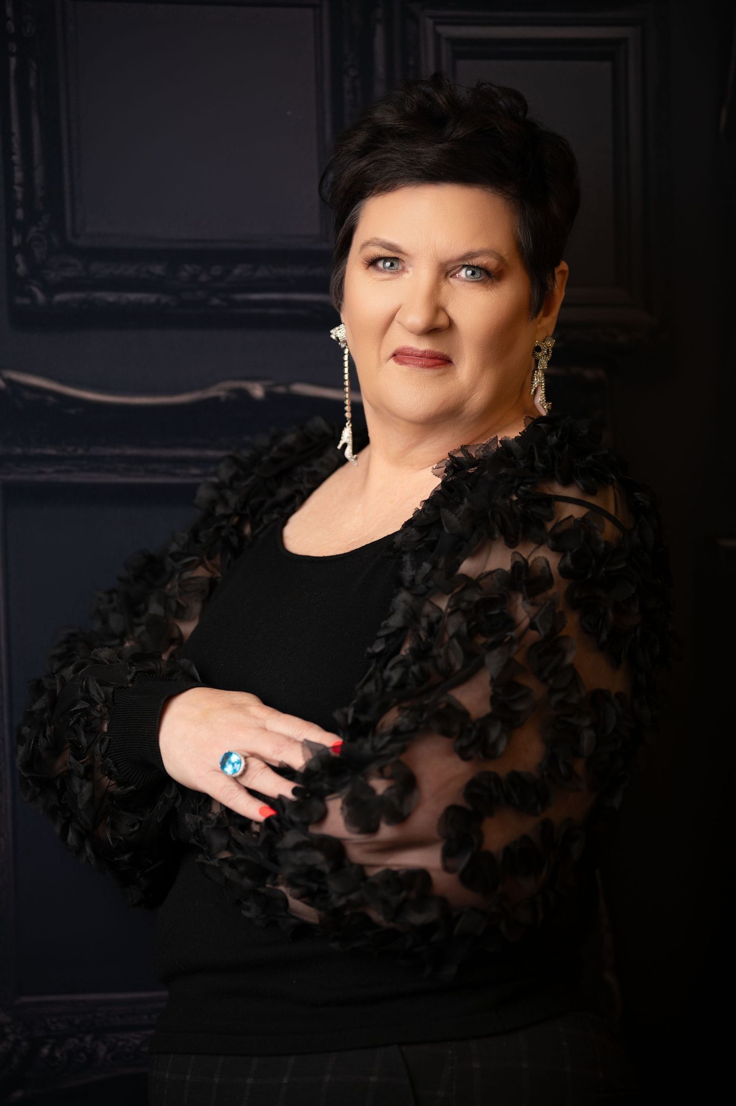 Studio portrait of person in black lace top with statement earrings against dark background.