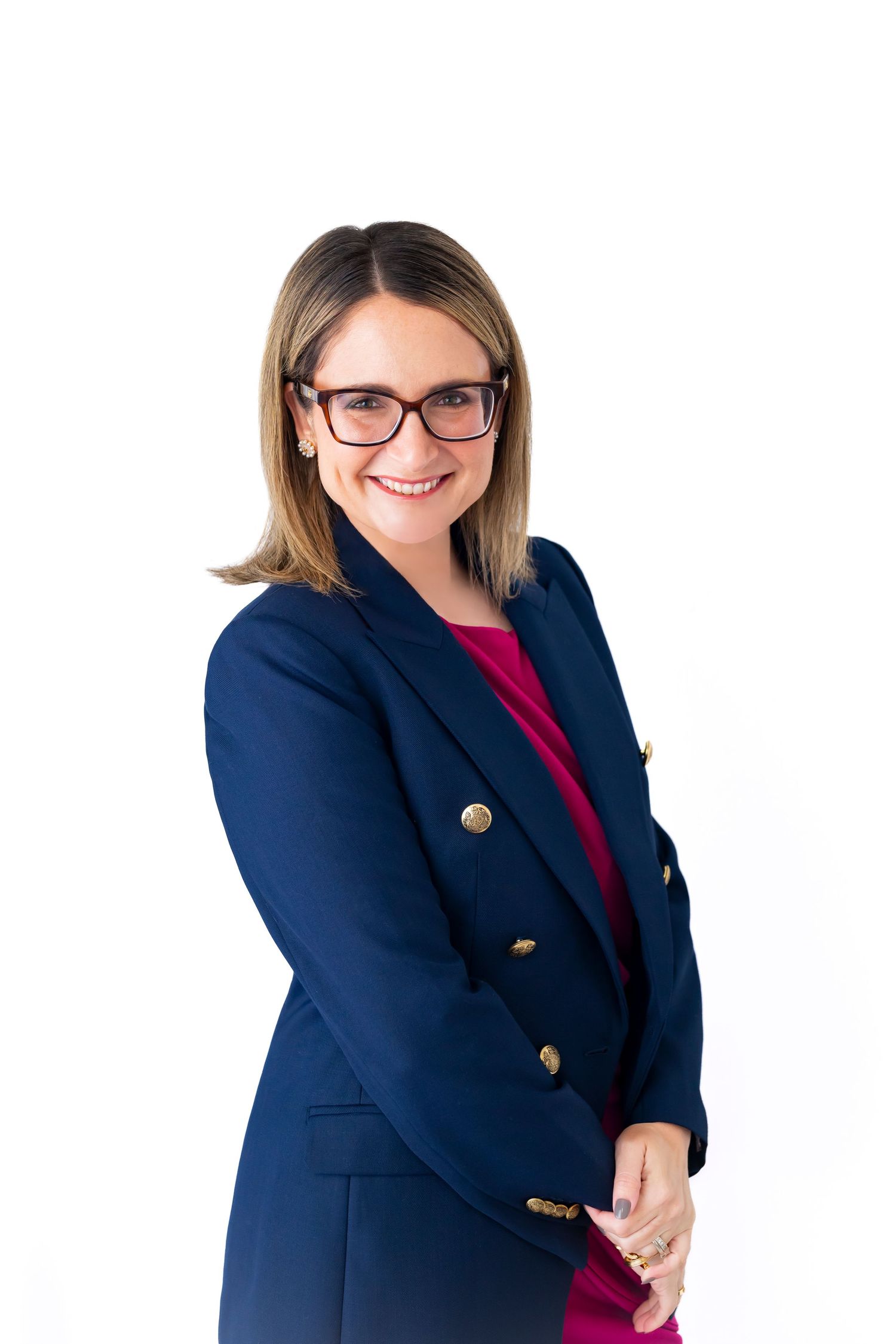 Professional headshot of someone wearing glasses and a navy blazer with gold buttons over a red dress.
