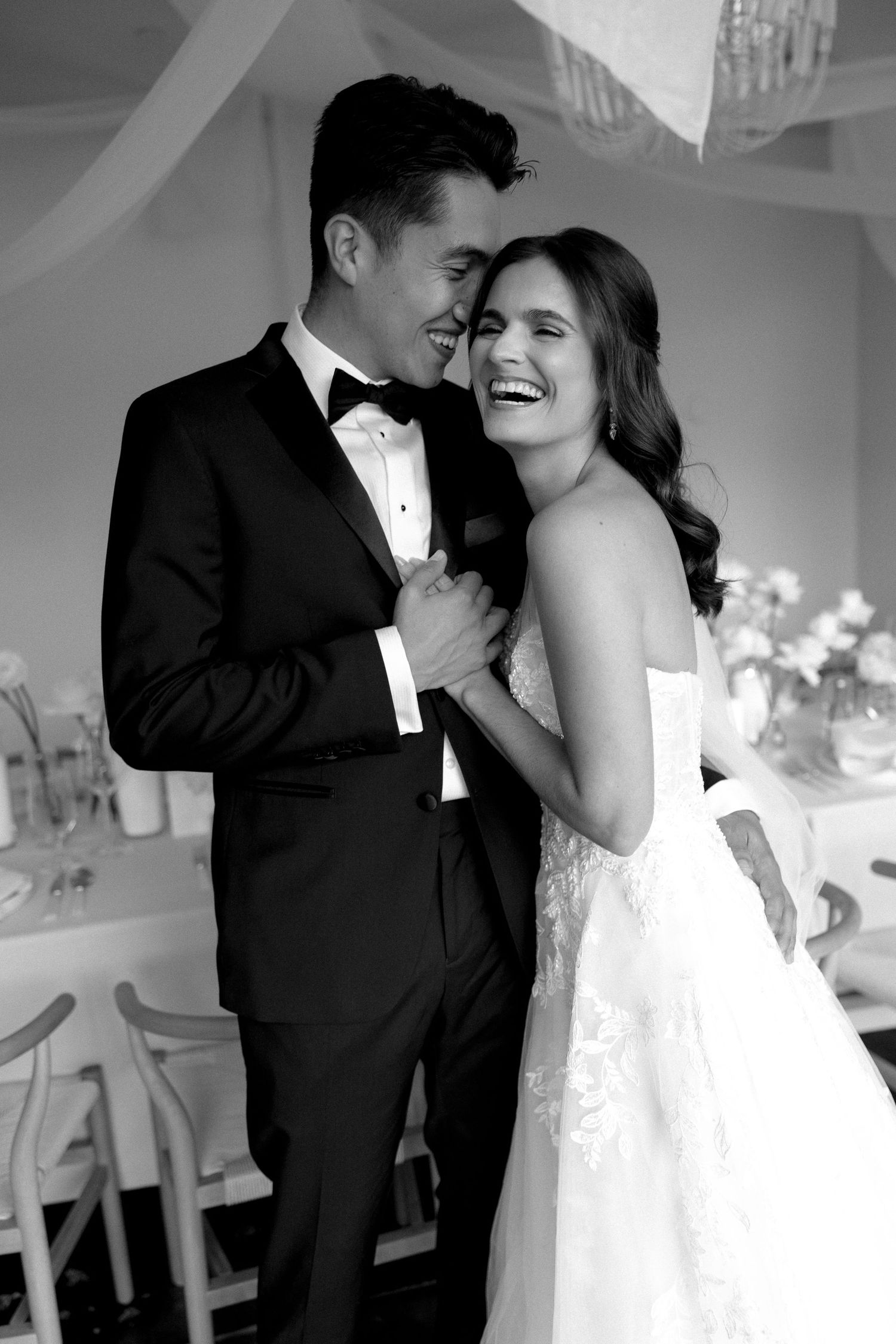 A black and white wedding photo showing a couple laughing together, with the bride in white and groom in tuxedo.