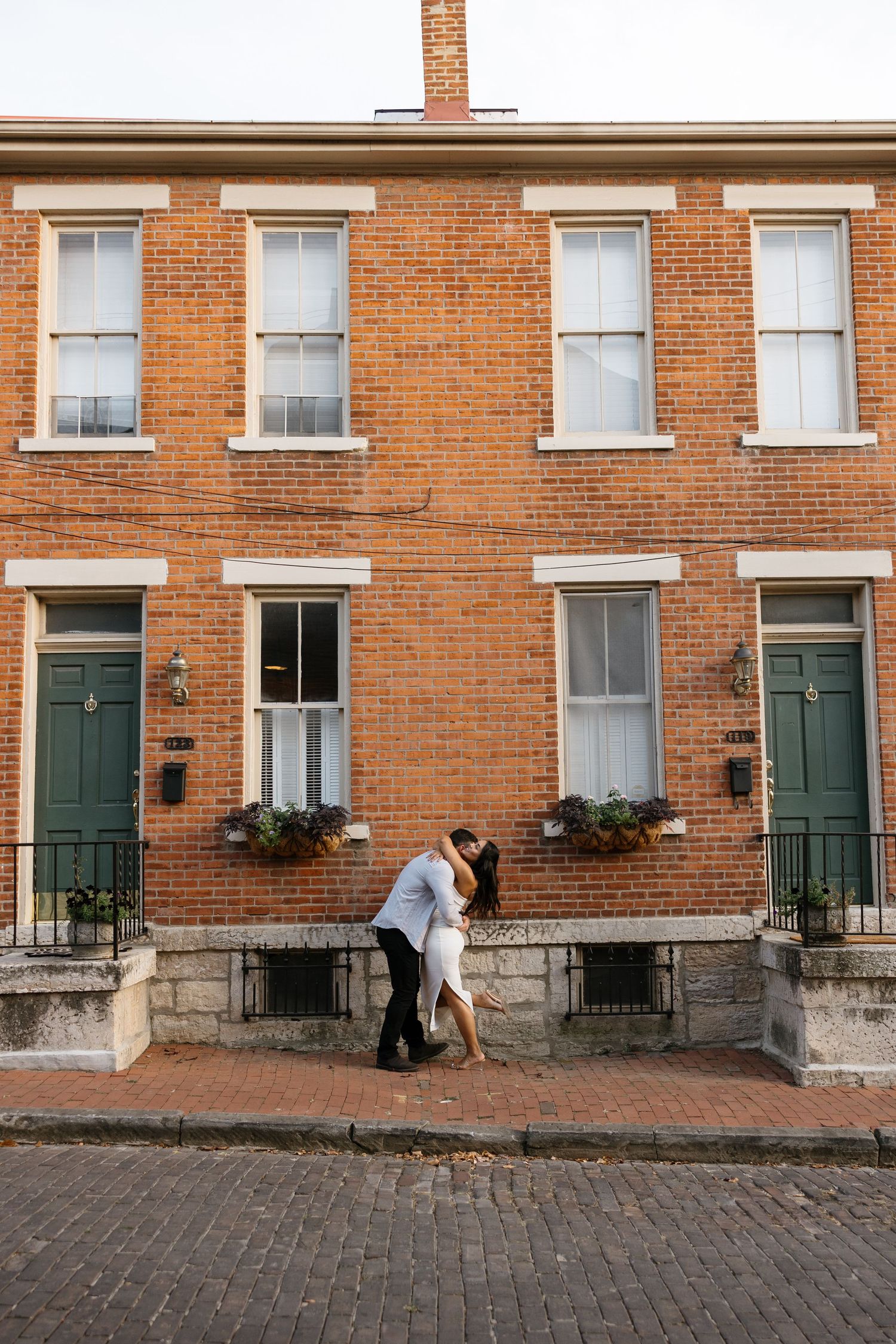 Couple sharing a romantic dip kiss on a brick cobblestone street in front of a charming red brick building with green doors.