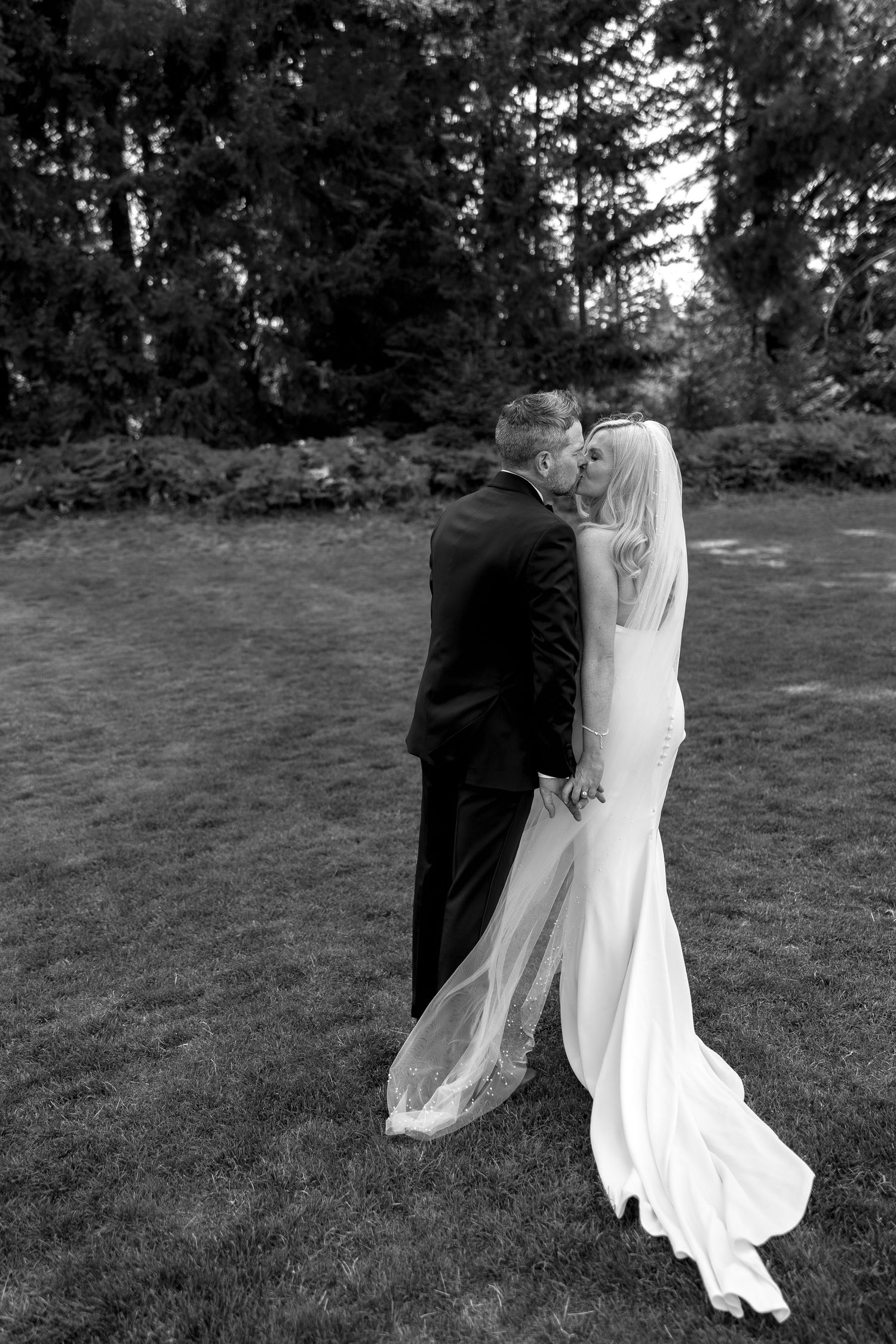 Black and white photo of bride and groom sharing a kiss outdoors on green lawn with trees in background.