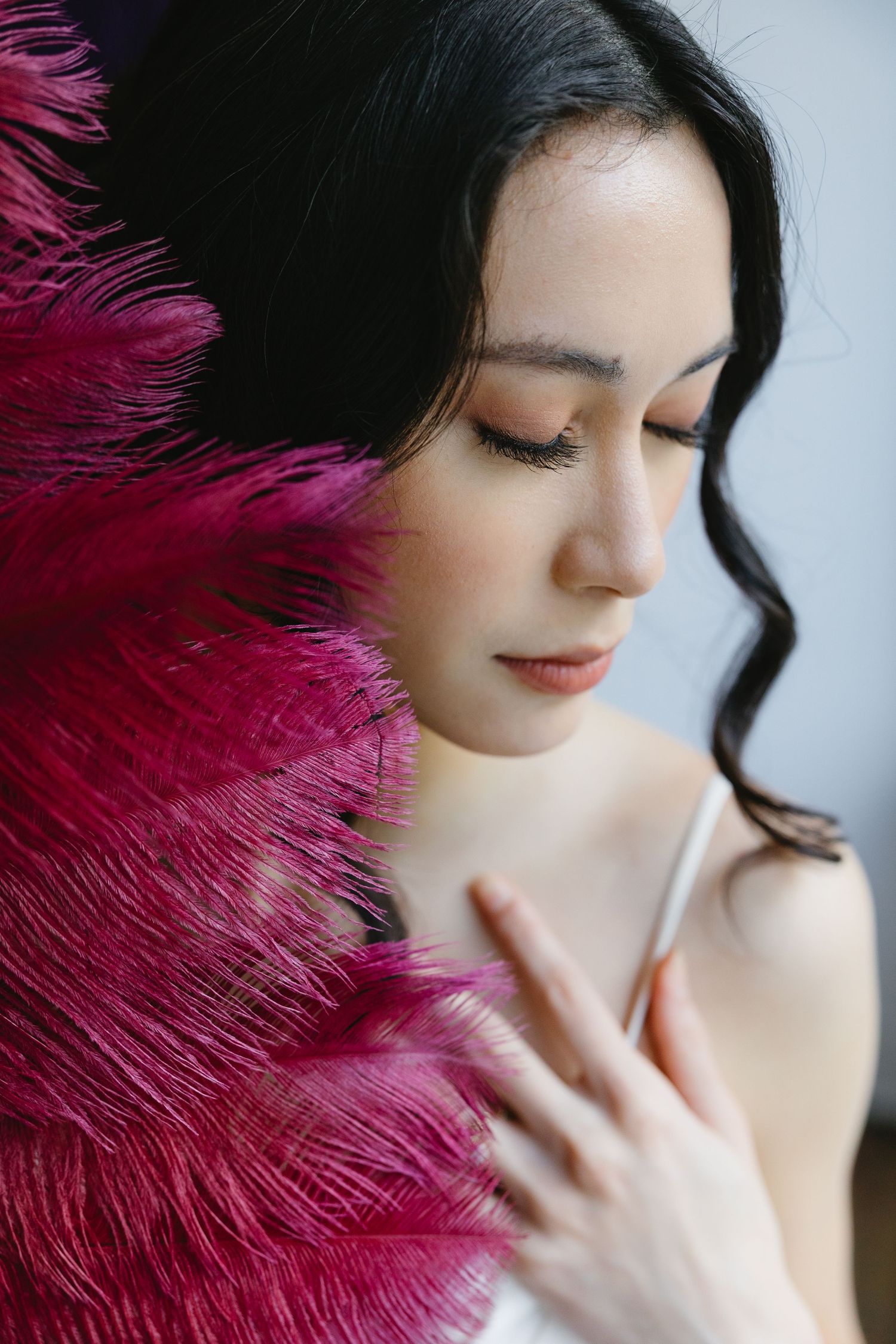 A young Asian woman with dark hair holds vibrant pink feathers, eyes closed, in an elegant and serene portrait.