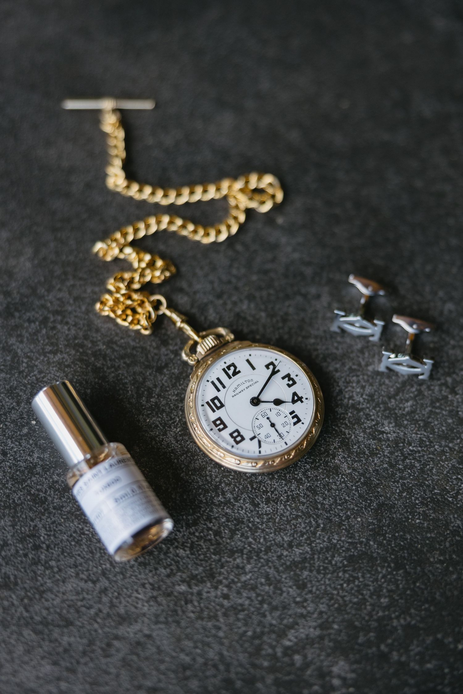 Vintage gold pocket watch with chain displayed alongside silver cufflinks and a small perfume bottle on dark background.