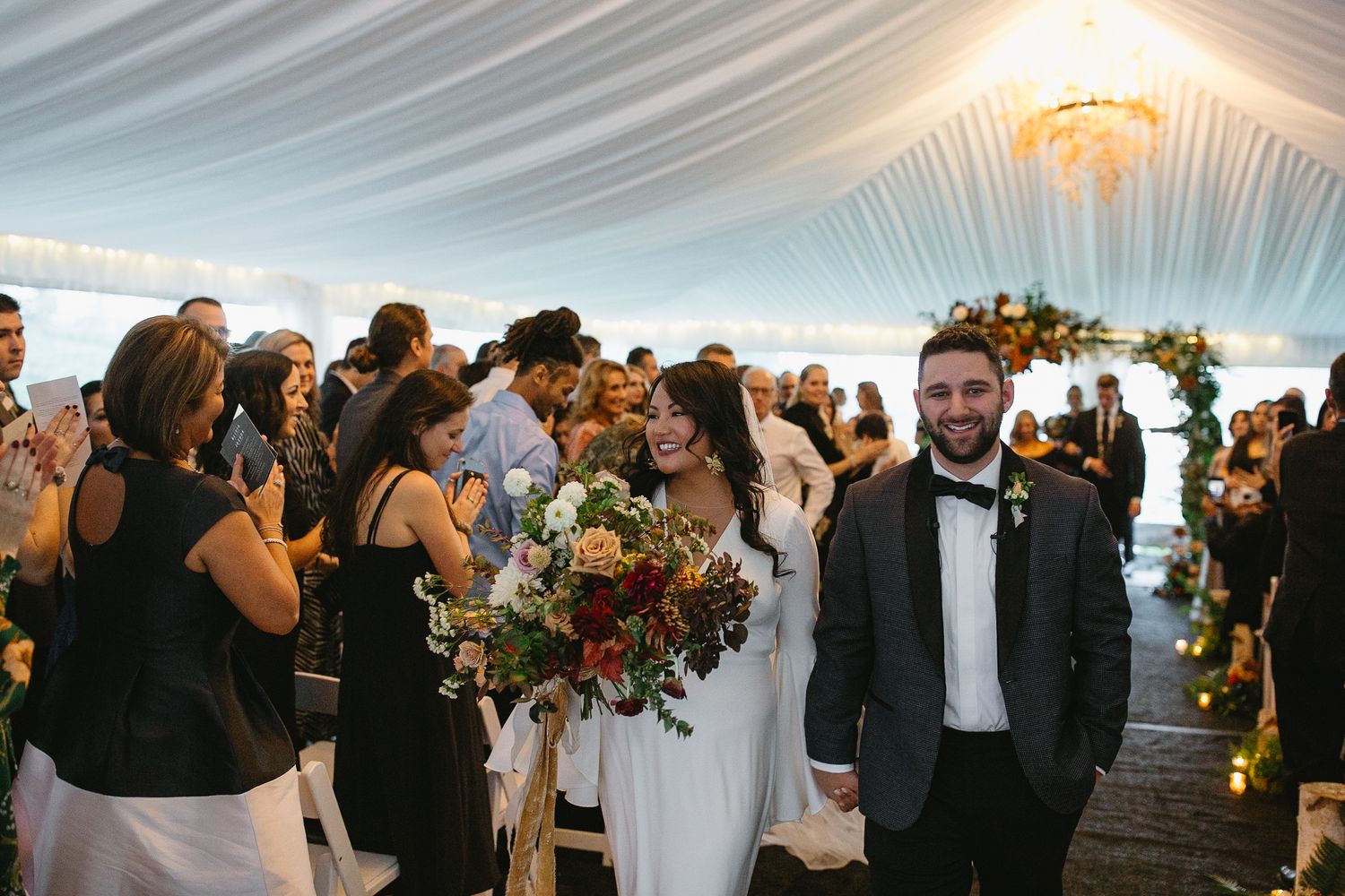 Newlywed couple walks down the aisle smiling, bride holding a lush bouquet, guests cheering in a draped tent venue.
