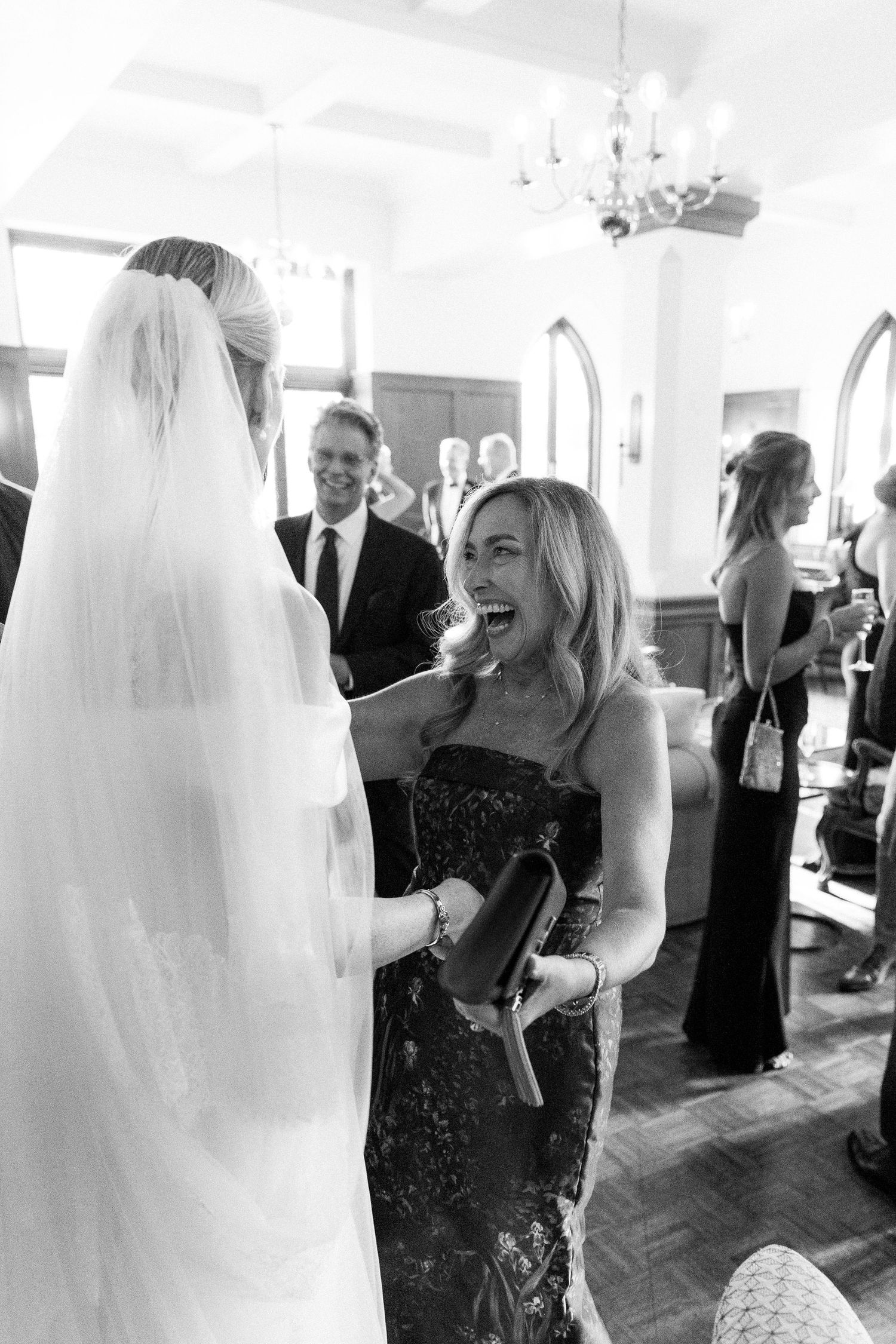 Black and white photo of a bride in veil laughing with a guest in a lace gown at an elegant wedding reception.