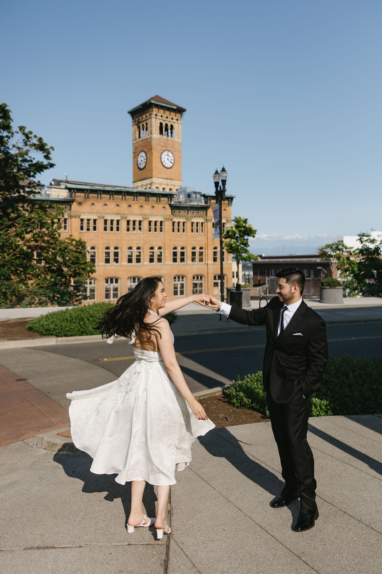 Bride and groom dancing outdoors near a historic clock tower building on a sunny day.
