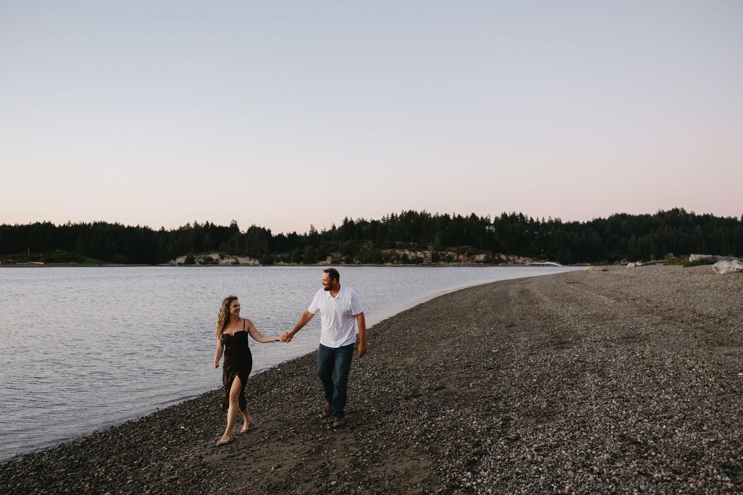 Couple holding hands walking along a rocky beach shoreline at dusk with evergreen trees in the background.