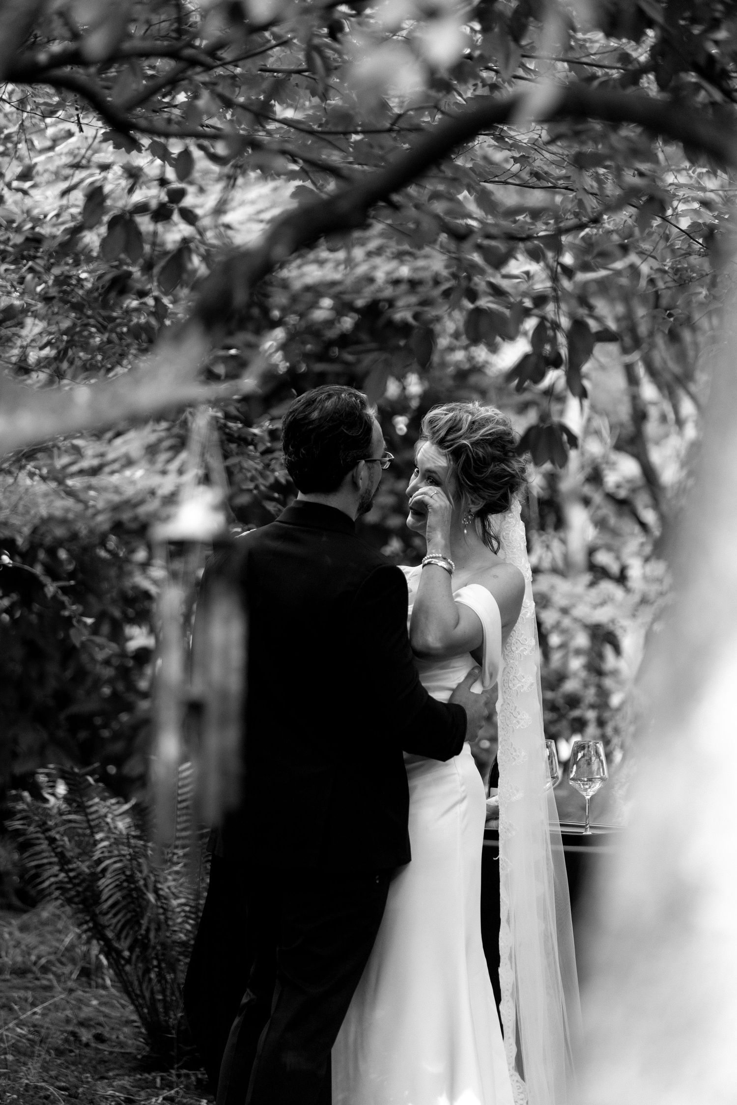A romantic black and white wedding ceremony scene captured through tree branches and leaves where the bride wipes tears.