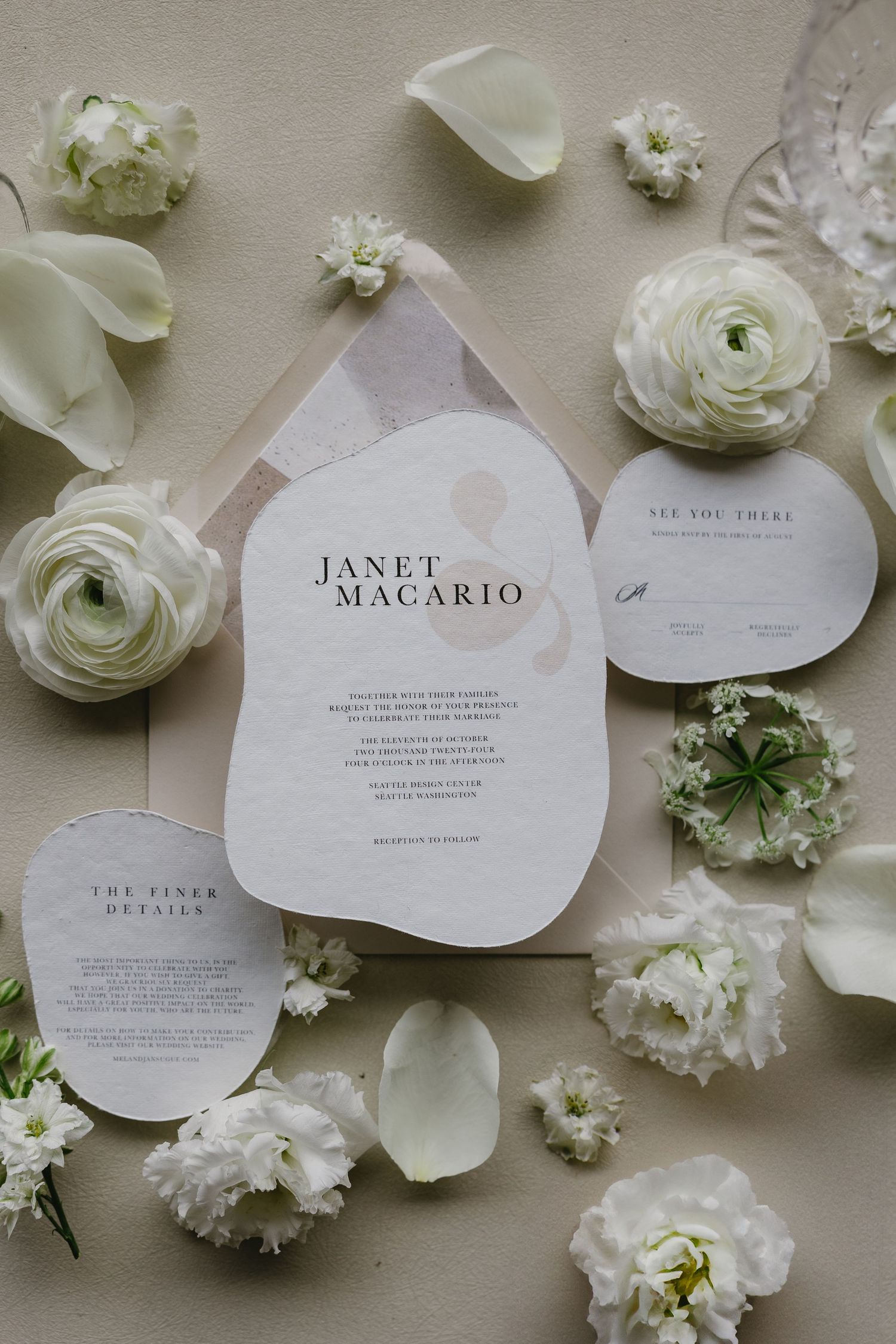 Elegant wedding invitation cards surrounded by delicate white ranunculus flowers and petals on a light beige surface.