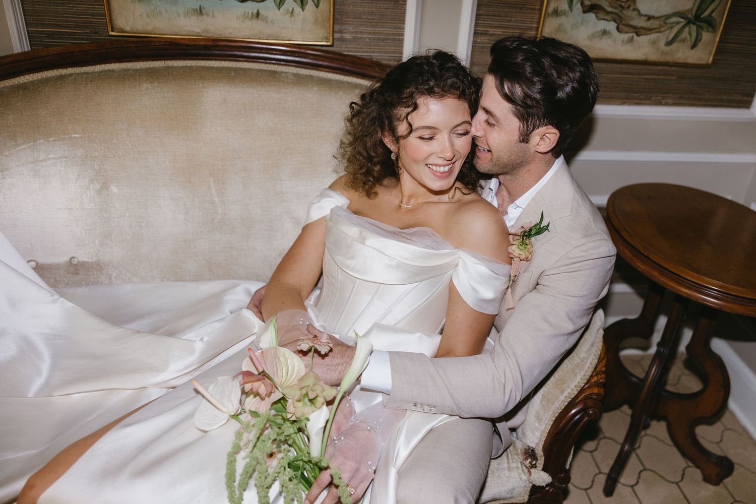 A couple in wedding attire share an intimate moment while seated on an elegant cream-colored sofa.