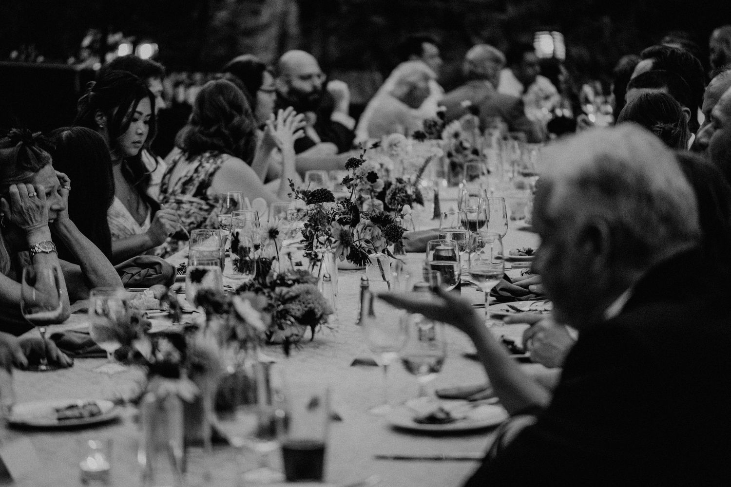 A black and white photograph of an elegant dinner party with guests seated at a long table with floral centerpieces.