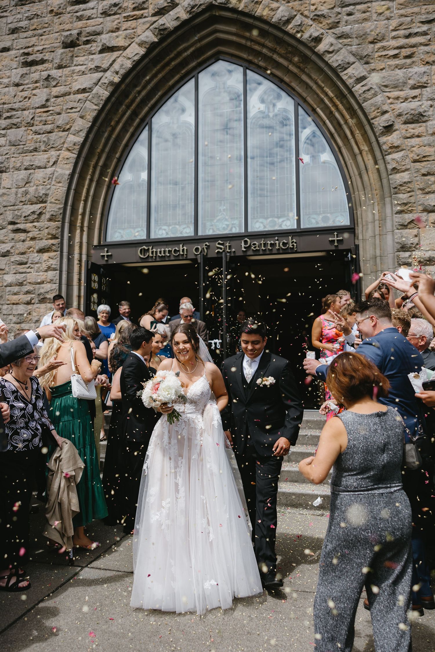 Newlyweds exit church under shower of confetti while guests celebrate outside stone archway of Guardian St. Patrick.