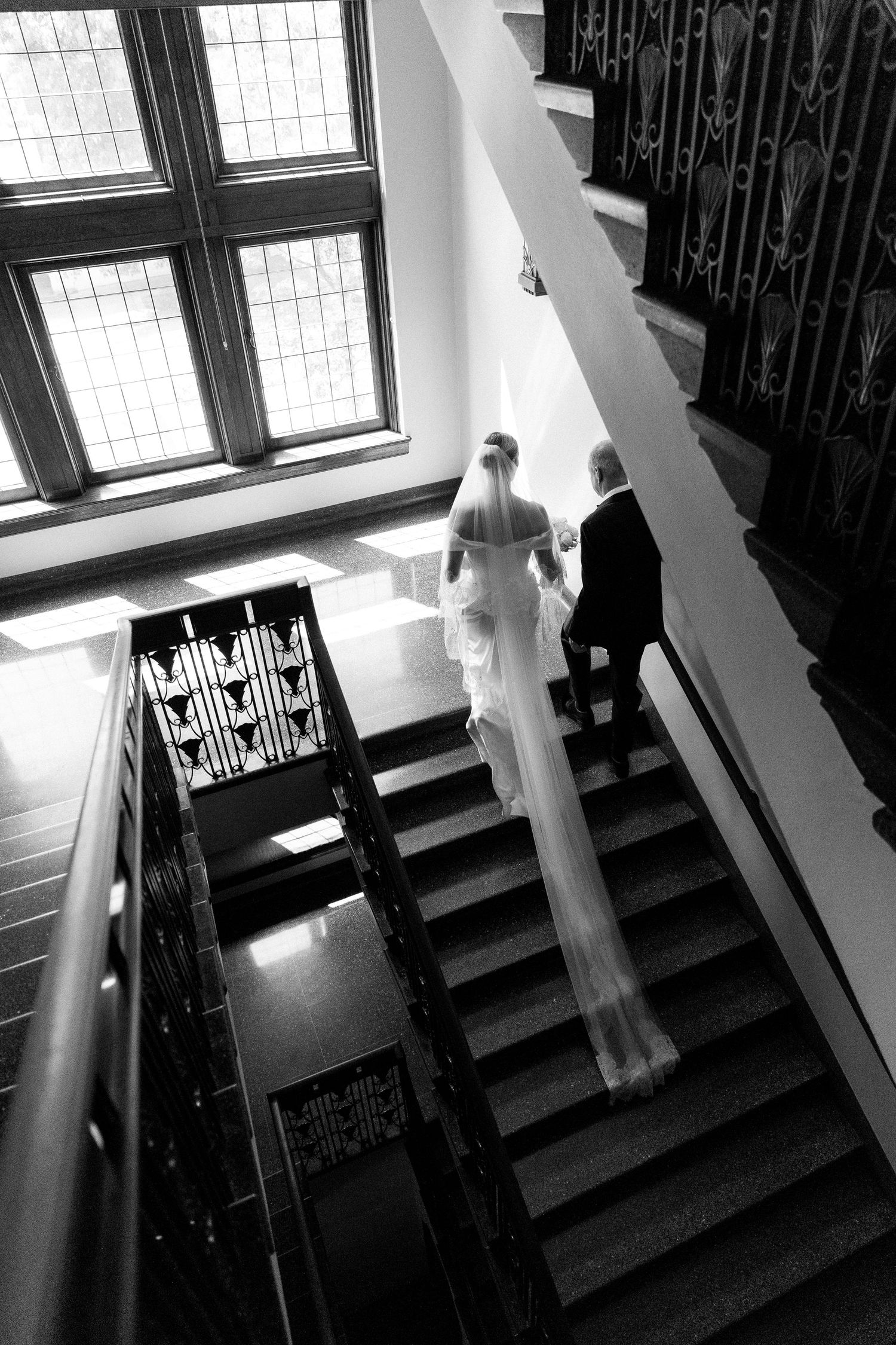 Black and white aerial view of a bride in a flowing veil ascending elegant stairs with a companion at a wedding venue.