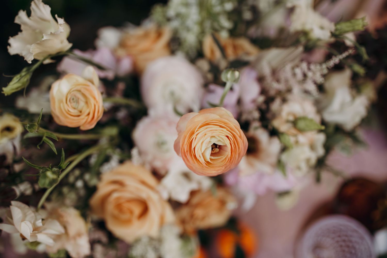 Delicate peach and pink floral arrangement featuring ranunculus and roses in soft pastel tones against a blurred background.