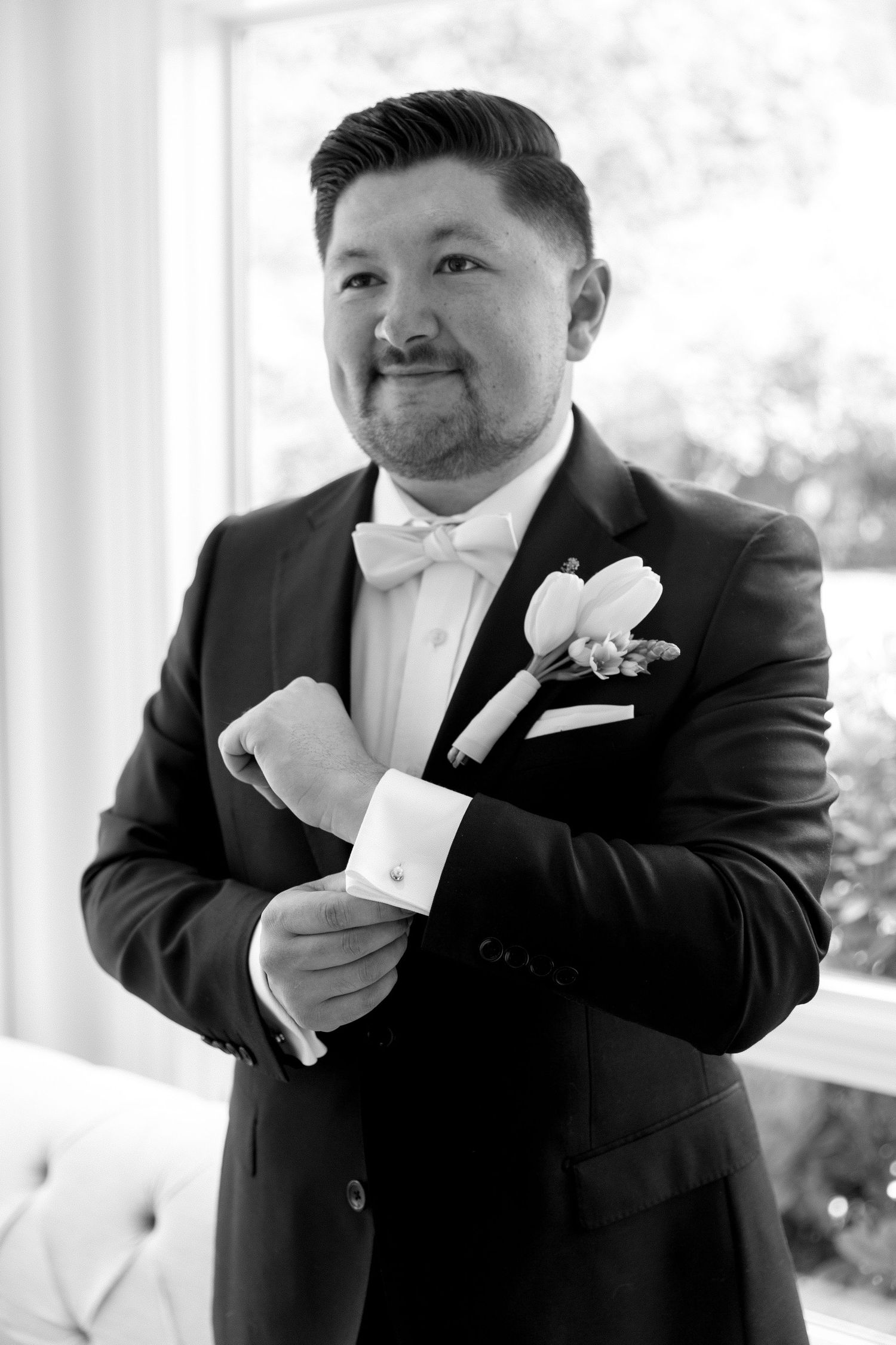 Black and white photo of a groom in a tuxedo with bow tie and boutonniere, adjusting his cufflinks before the wedding.