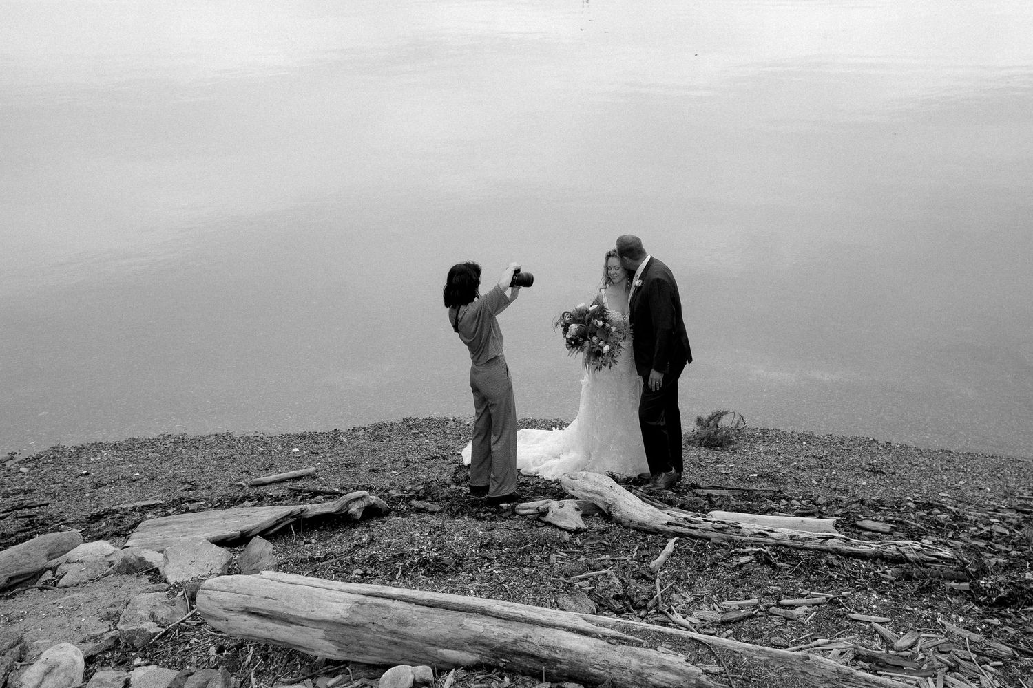 Black and white photo of a couple sharing a kiss at their outdoor wedding ceremony near a misty lake.