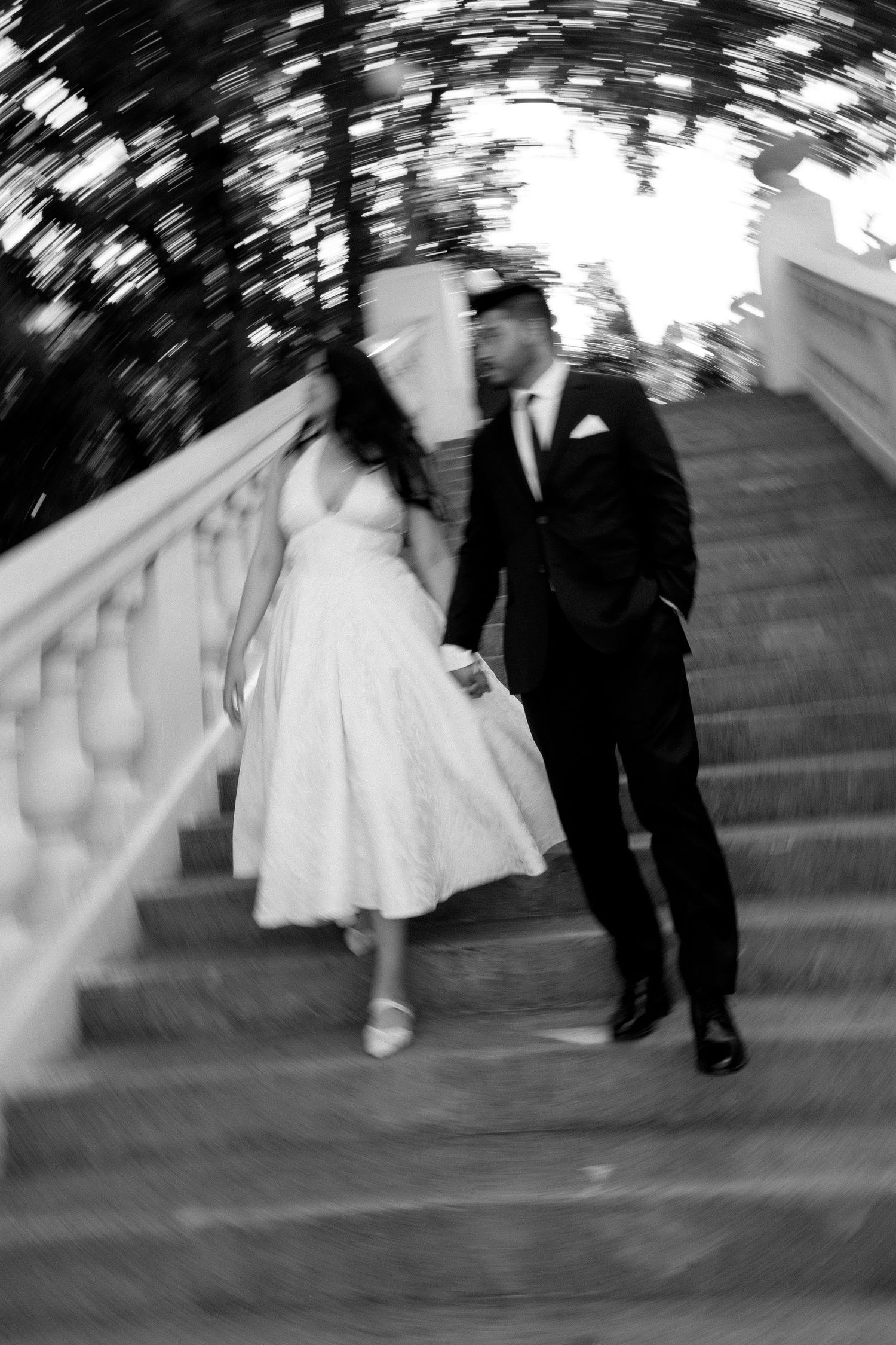 Black and white photo of bride and groom holding hands walking up elegant stone staircase with balustrade.