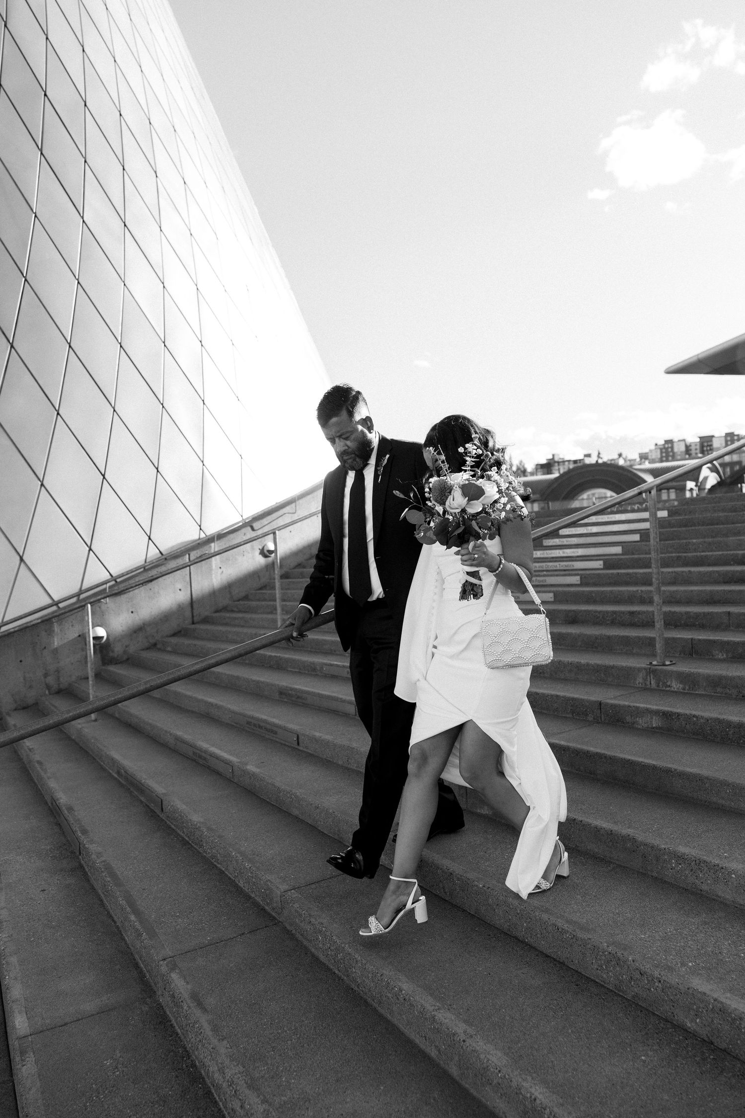 Black and white photo of a bride and groom walking down outdoor stairs, bride holding bouquet, modern building backdrop.