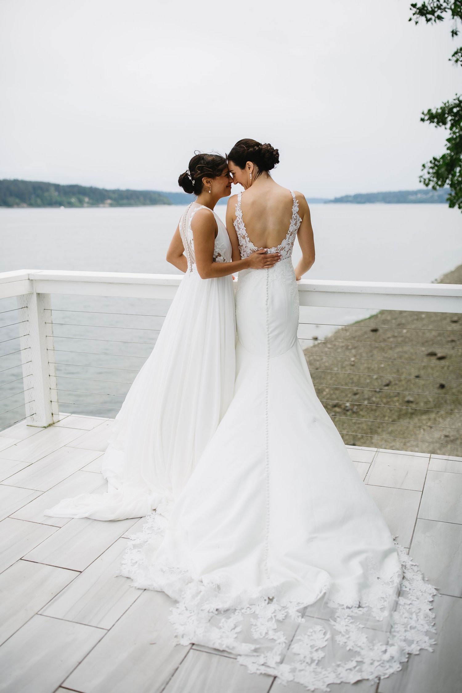 Two brides embrace on a white stone terrace overlooking a scenic waterfront view.