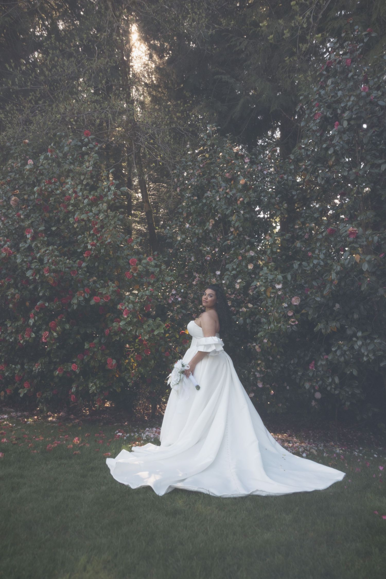 Bride in elegant off-shoulder white ball gown with long train stands in lush garden surrounded by blooming trees.