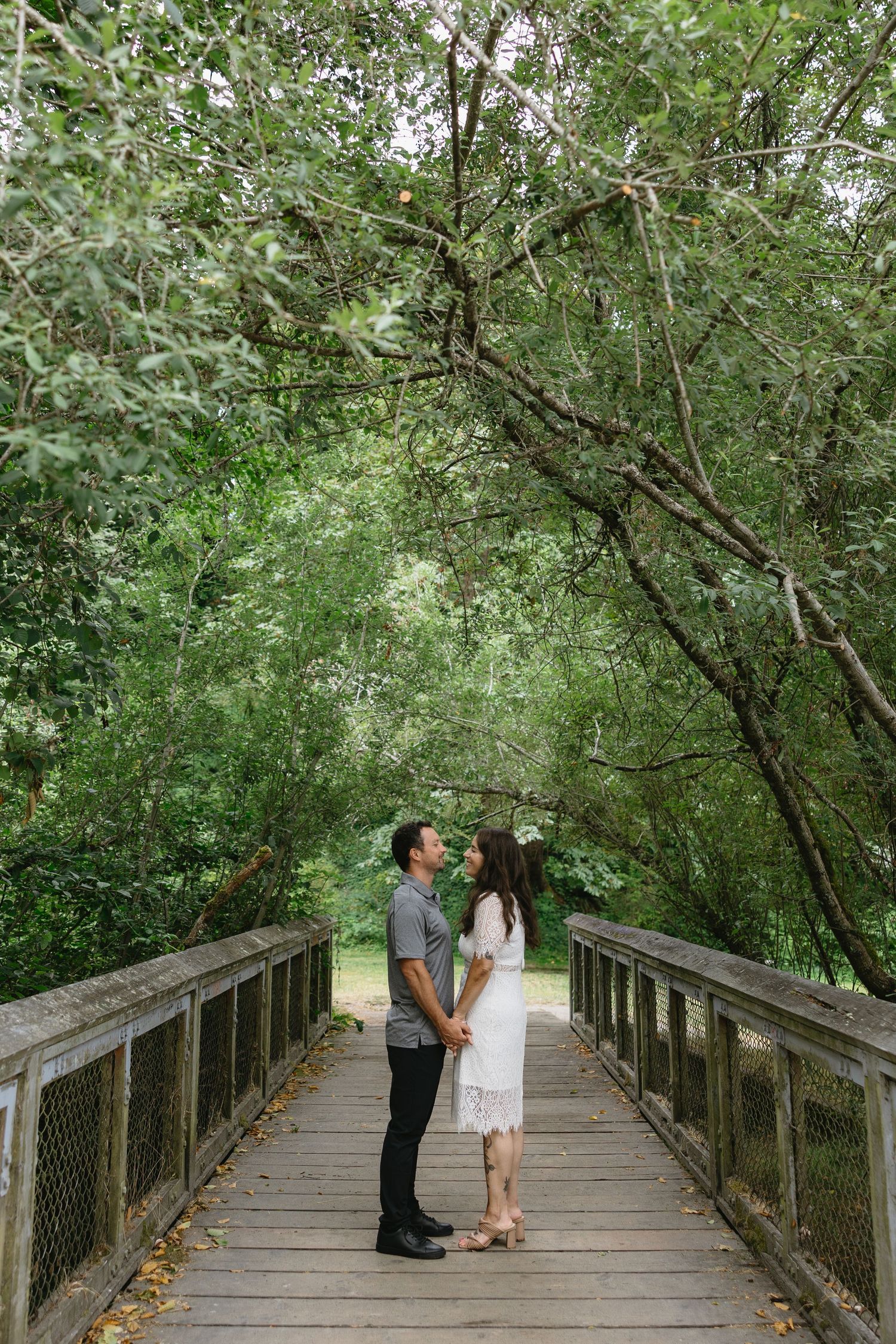 Couple holding hands facing each other on a wooden bridge surrounded by lush green trees in a park.