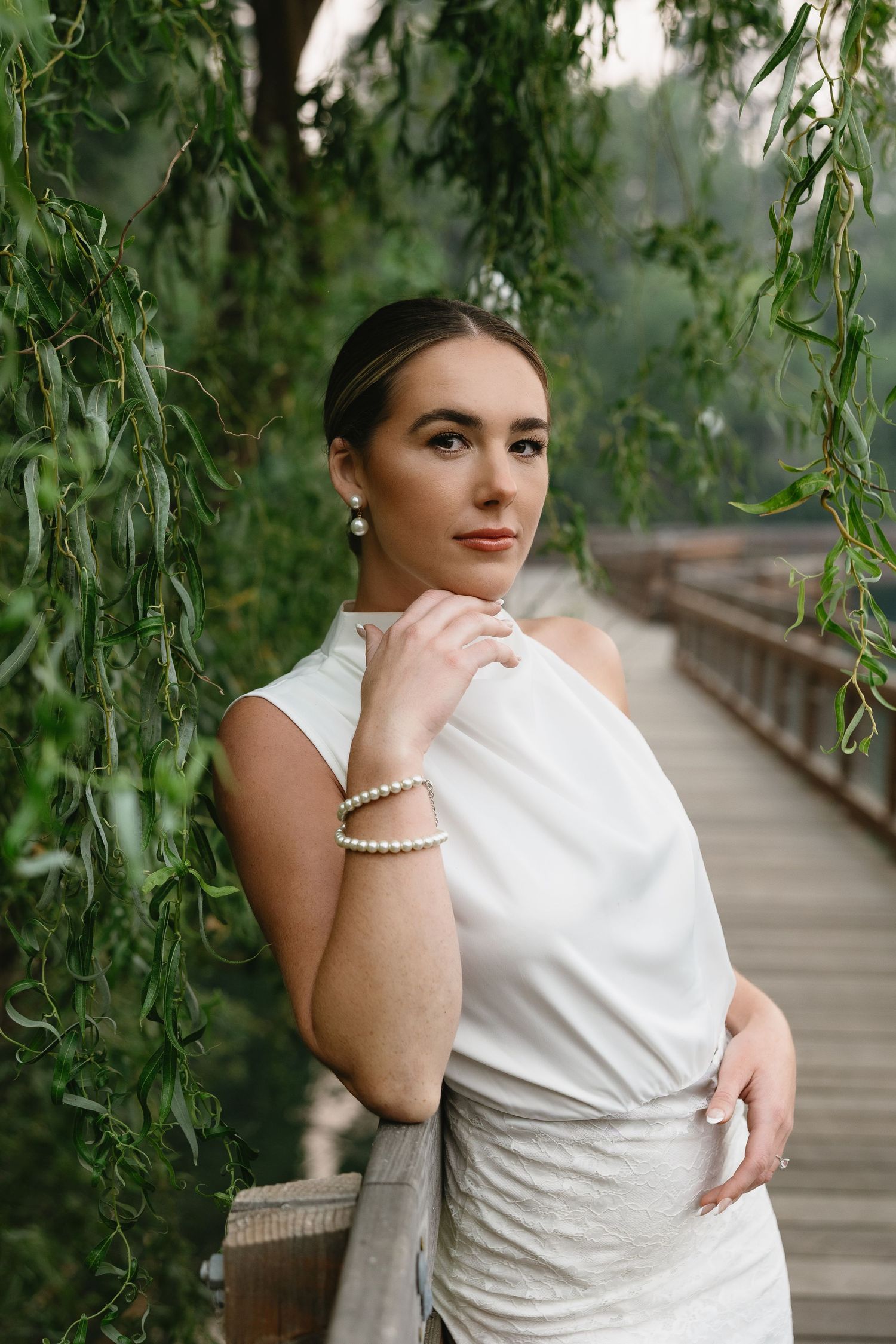 Elegant woman in white dress with pearl bracelet posing on wooden bridge surrounded by green willow trees.