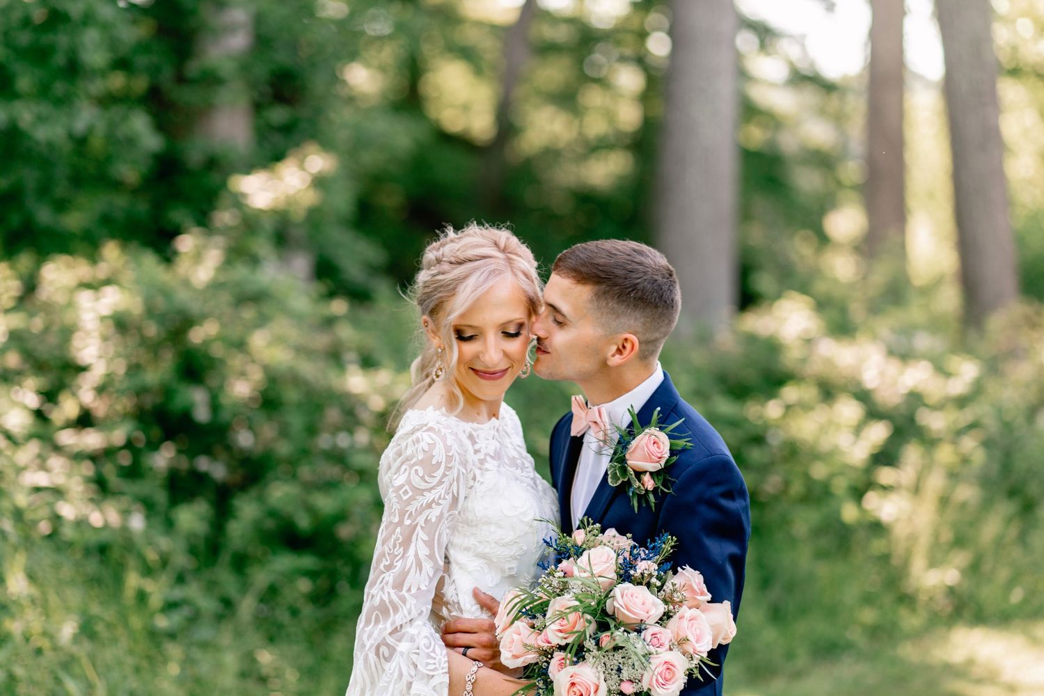 Bride and groom share a romantic moment outdoors, surrounded by lush green trees, as groom kisses bride holding pink bouquet.