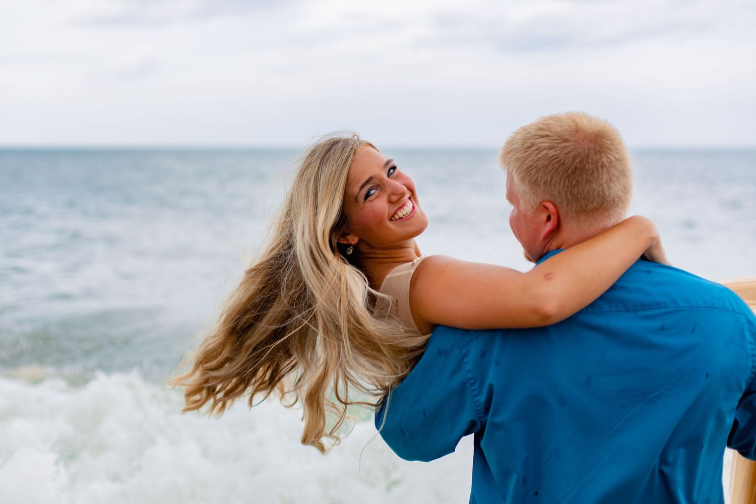 Joyful woman with flowing blonde hair embraces her partner on a beach with waves crashing behind them on a cloudy day.