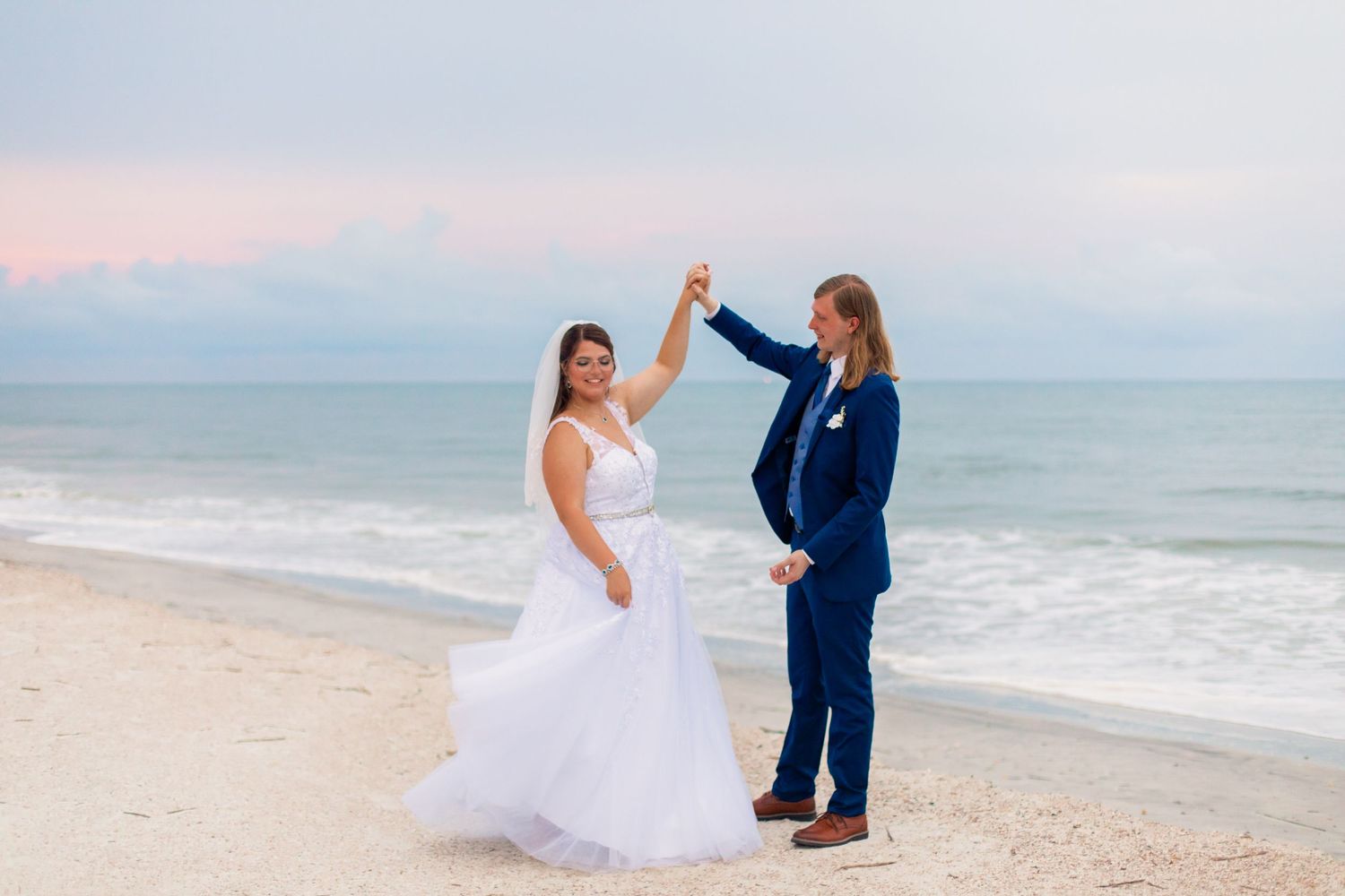 Bride in white gown and groom in navy suit dance on a sandy beach at sunset with pastel pink and blue skies.