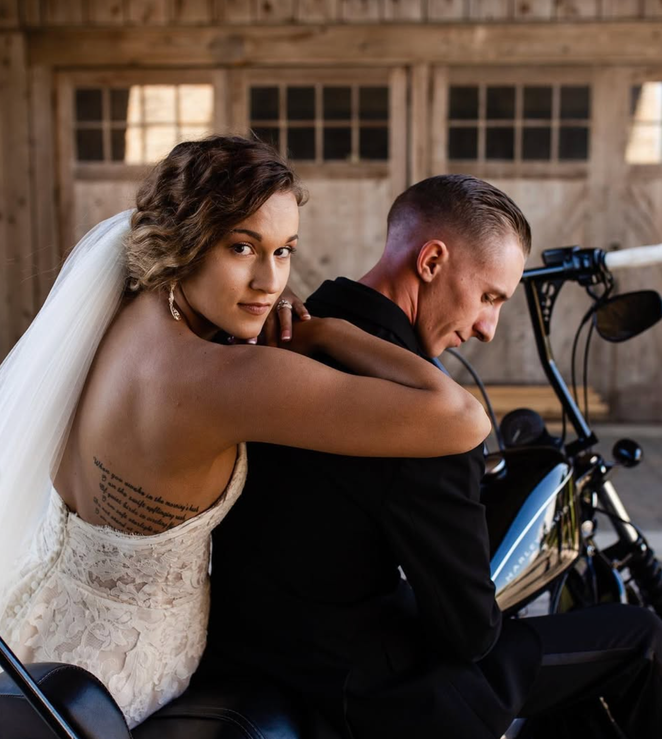 Bride and groom pose on motorcycle inside rustic wooden barn on wedding day.