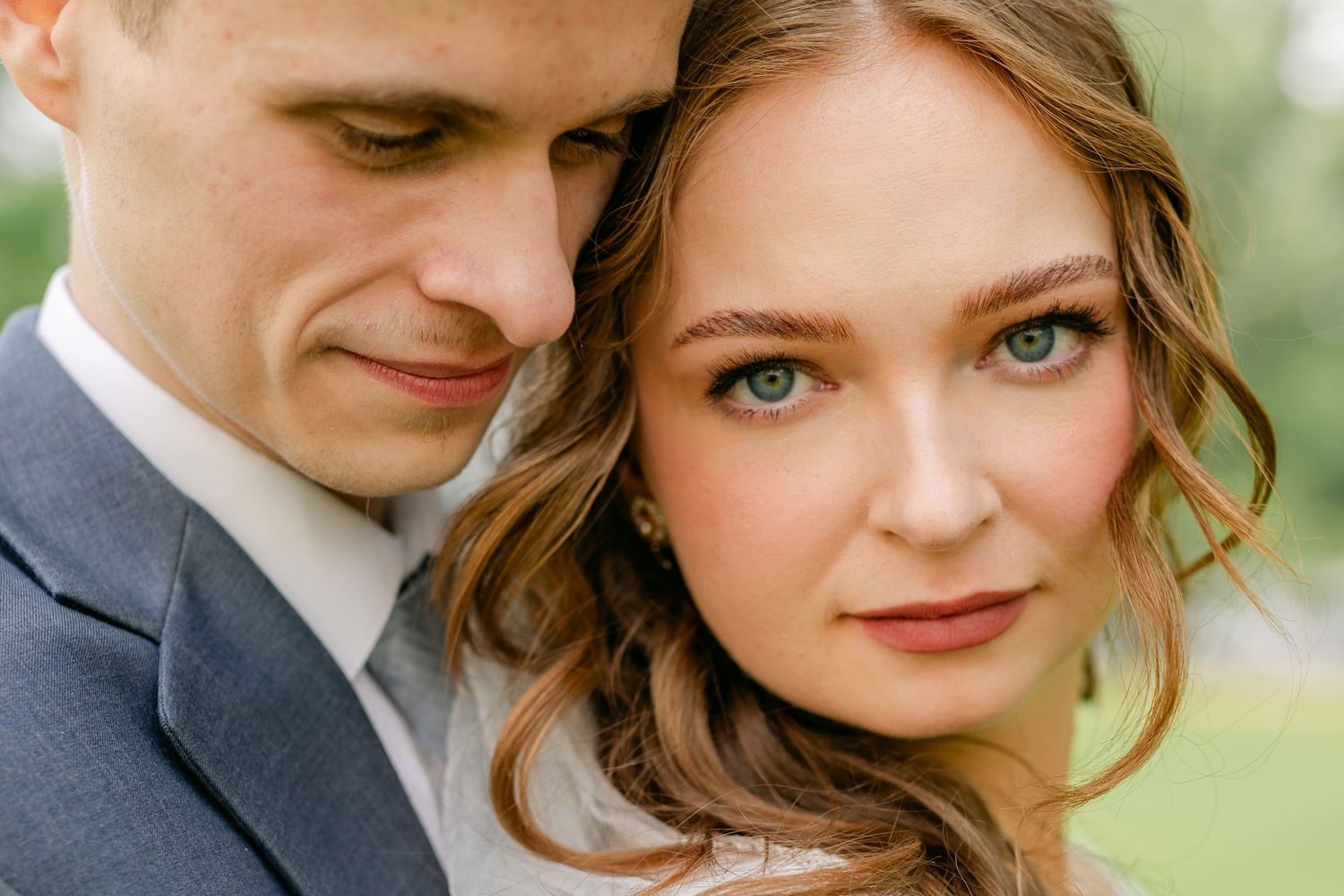Close-up portrait of bride with blue eyes leaning on groom in gray suit outdoors.