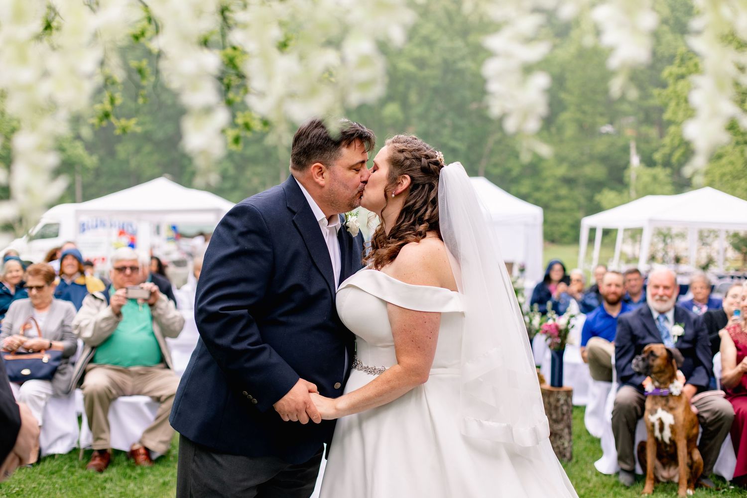 Bride and groom sharing their first kiss at an outdoor wedding ceremony under white floral arrangements.