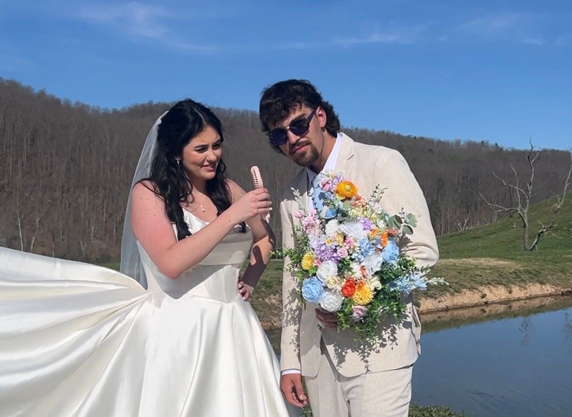 Bride and groom stand by lakeside holding colorful bouquet under bright blue sky.