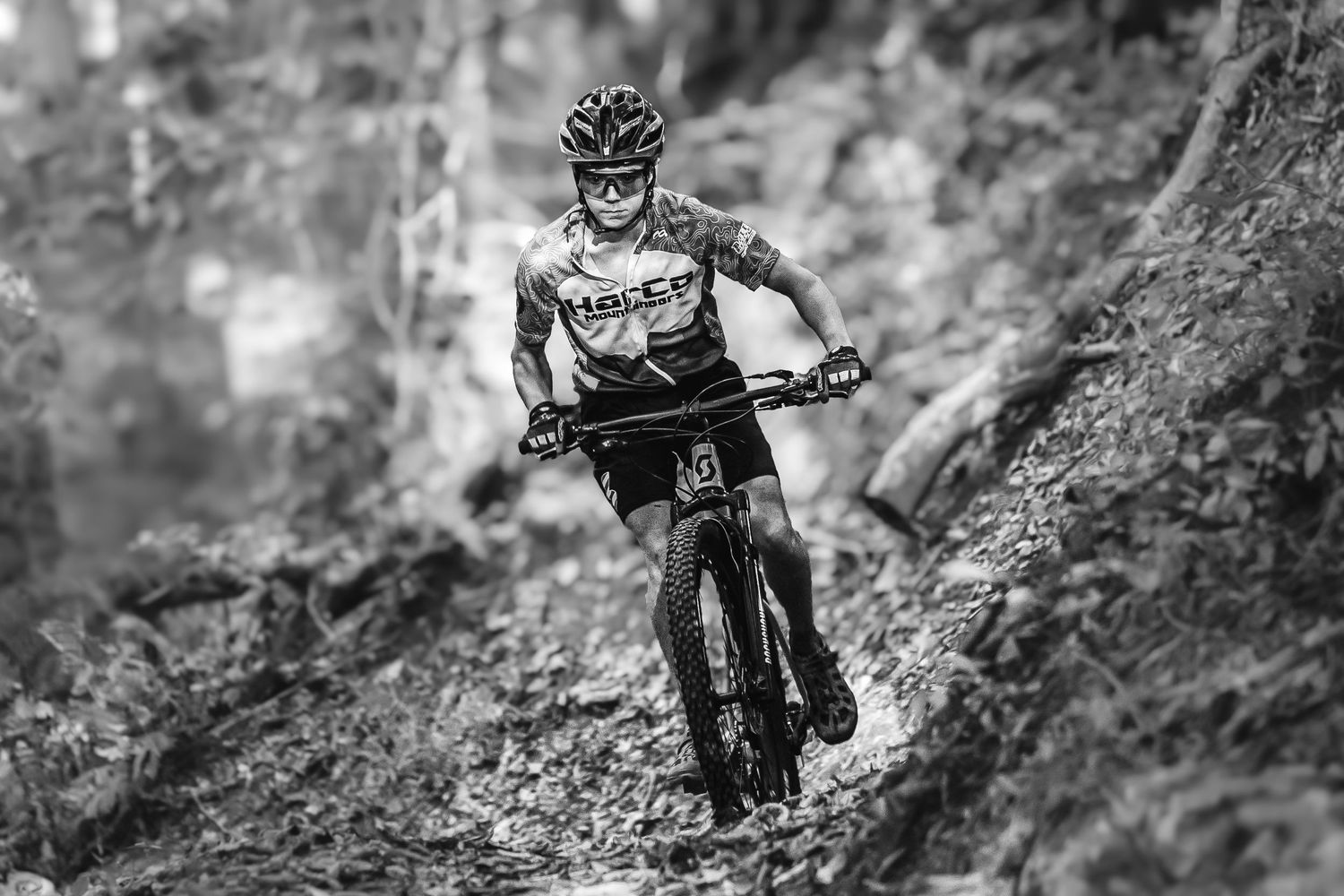 Black and white photo of a mountain biker racing downhill on a rocky trail wearing a helmet and racing jersey.