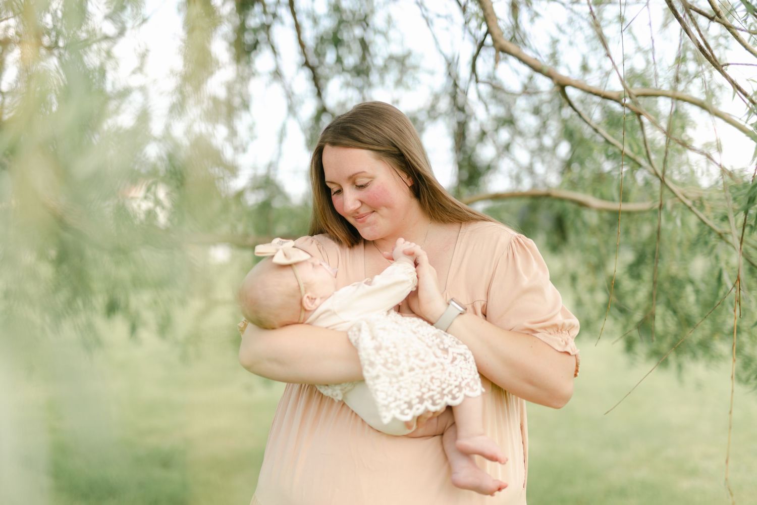 Mother tenderly cradling and nursing newborn baby outdoors beneath weeping willow tree branches in soft natural light.